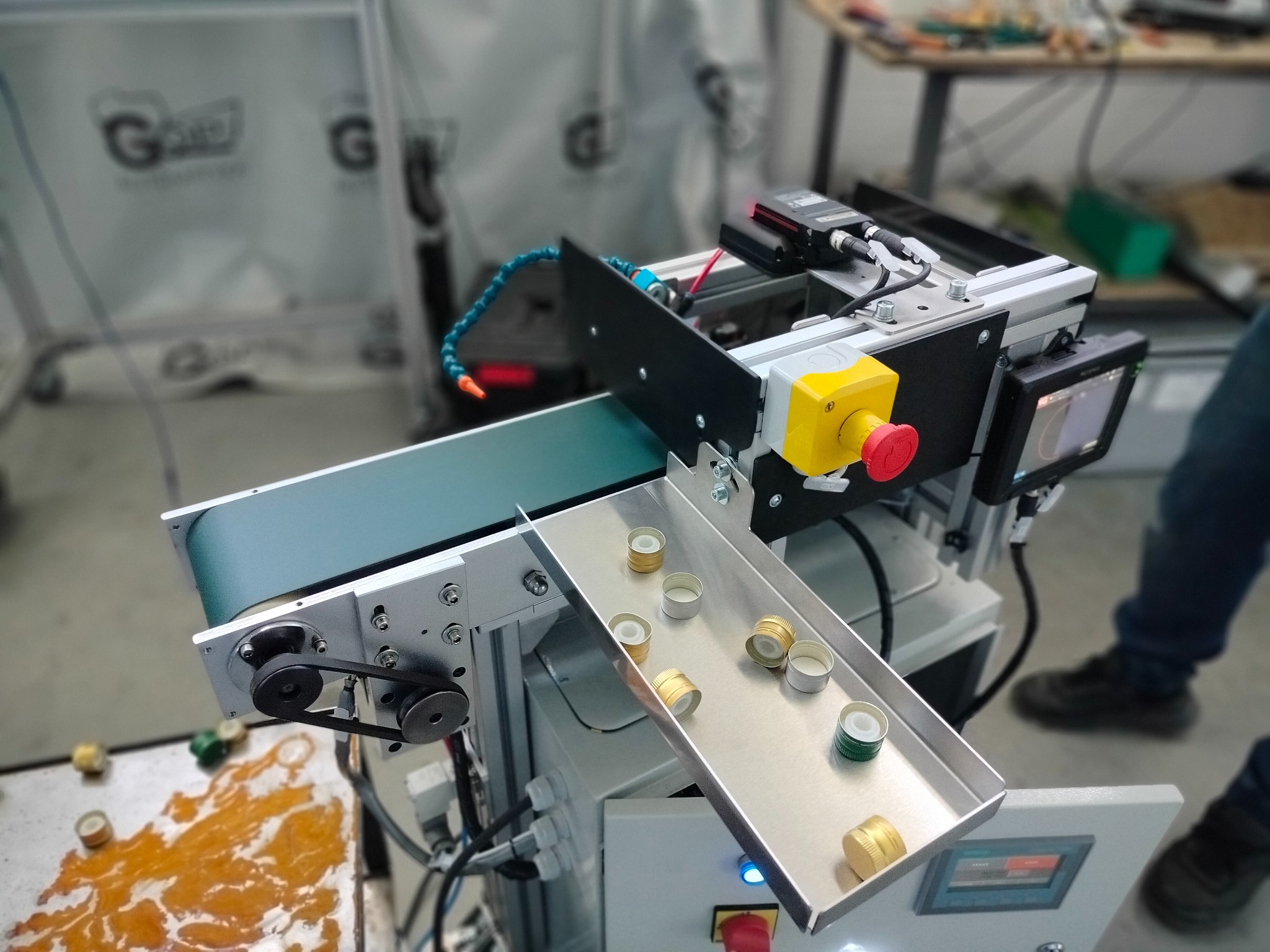 Automatyczna linia produkcyjna z taśmociągiem sortująca metalowe nakrętki, widoczny panel sterowania i czujniki. W tle zamazane logo firmy.