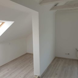 Kwadrat - Poddasze po remoncie: świeżo pomalowane białe ściany, drewniane belki stropowe, okno dachowe, grzejnik pod oknem z niebieską folią ochronną, panele podłogowe imitujące drewno.