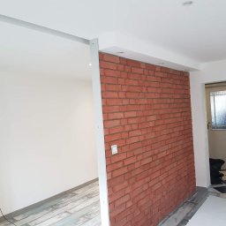 Kwadrat - Ściana z cegły dekoracyjnej w odnowionym wnętrzu, widoczne fragmenty podłogi z paneli i drzwi wejściowe.