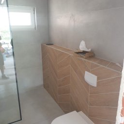 Kwadrat - Nowoczesna łazienka w trakcie wykańczania: podwieszana toaleta, drewnopodobne płytki ułożone w jodełkę na zabudowie stelaża, szklana kabina prysznicowa z czarnymi profilami, szare płytki...