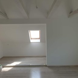 Kwadrat - Poddasze po remoncie z drewnianymi belkami stropowymi, jasnymi panelami podłogowymi i oknem dachowym, widoczna poziomica i pojemnik z farbą.