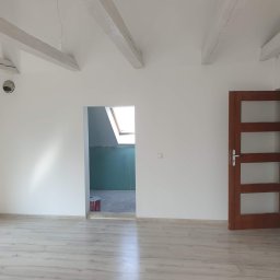 Kwadrat - Poddasze po remoncie: białe ściany, drewniane belki stropowe, jasna podłoga z paneli, widoczne drzwi wewnętrzne z przeszkleniami i fragment pomieszczenia z oknem dachowym i niepomalowanymi ścianami.