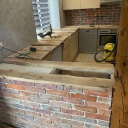 Kwadrat - Wnętrze kuchni w trakcie montażu drewnianego blatu z surowego drewna, widoczny mur z cegły, narzędzia, butelka oraz odkurzacz przemysłowy.