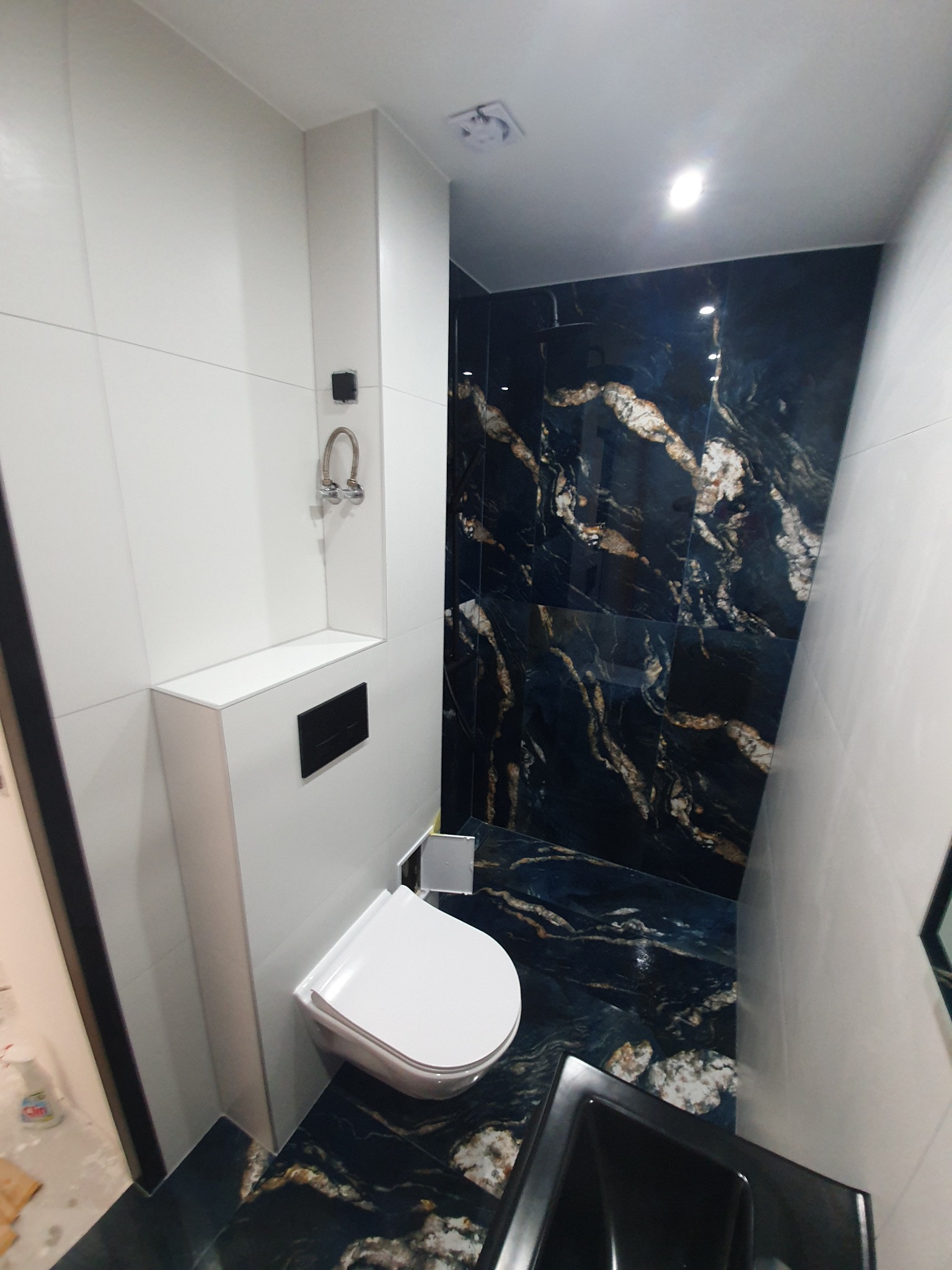 Wąska łazienka po remoncie z toaletą podwieszaną, biało-czarnymi płytkami imitującymi marmur i ukrytą spłuczką.