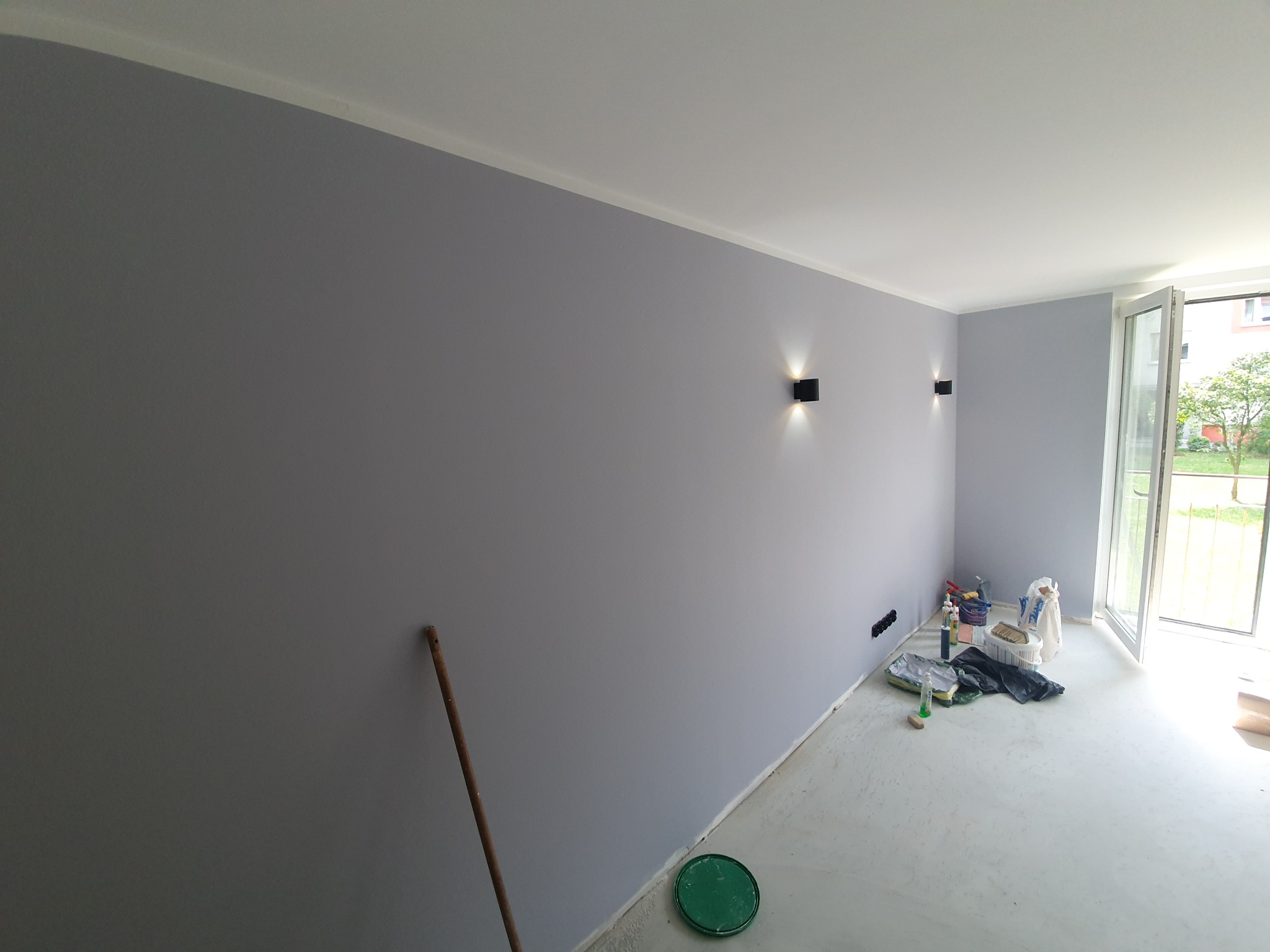 Świeżo pomalowane na szaro wnętrze z dwoma kinkietami, widoczne akcesoria malarskie i otwarte drzwi balkonowe wpadające światło dzienne.