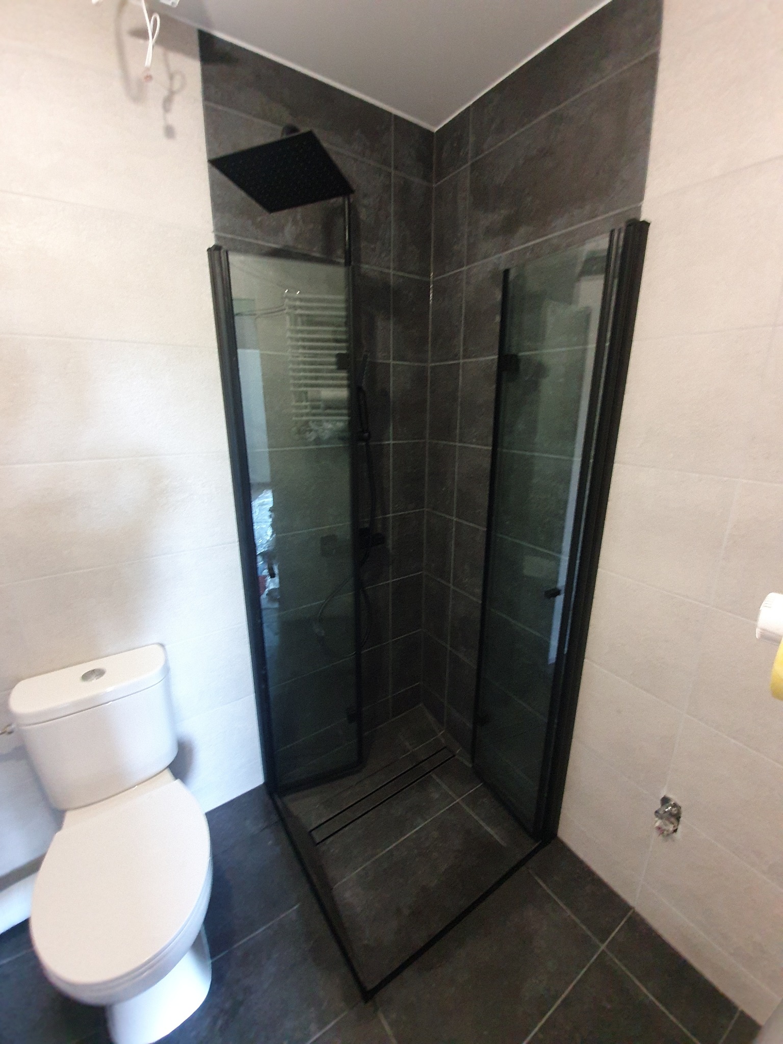 Nowoczesna łazienka z kabiną prysznicową typu walk-in z czarnymi ramami, deszczownicą i płytkami w odcieniach szarości, widoczna toaleta.