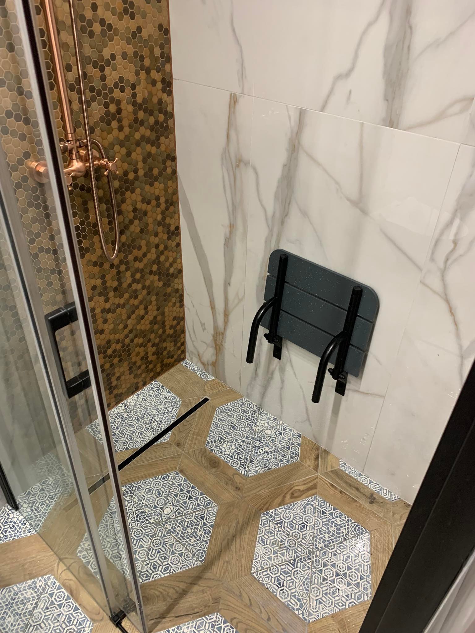 Nowoczesna kabina prysznicowa z mozaiką heksagonalną w odcieniach brązu, marmurowymi płytkami ściennymi, drewnopodobną podłogą w kształcie heksagonów z niebieskim wzorem i składanym siedziskiem.