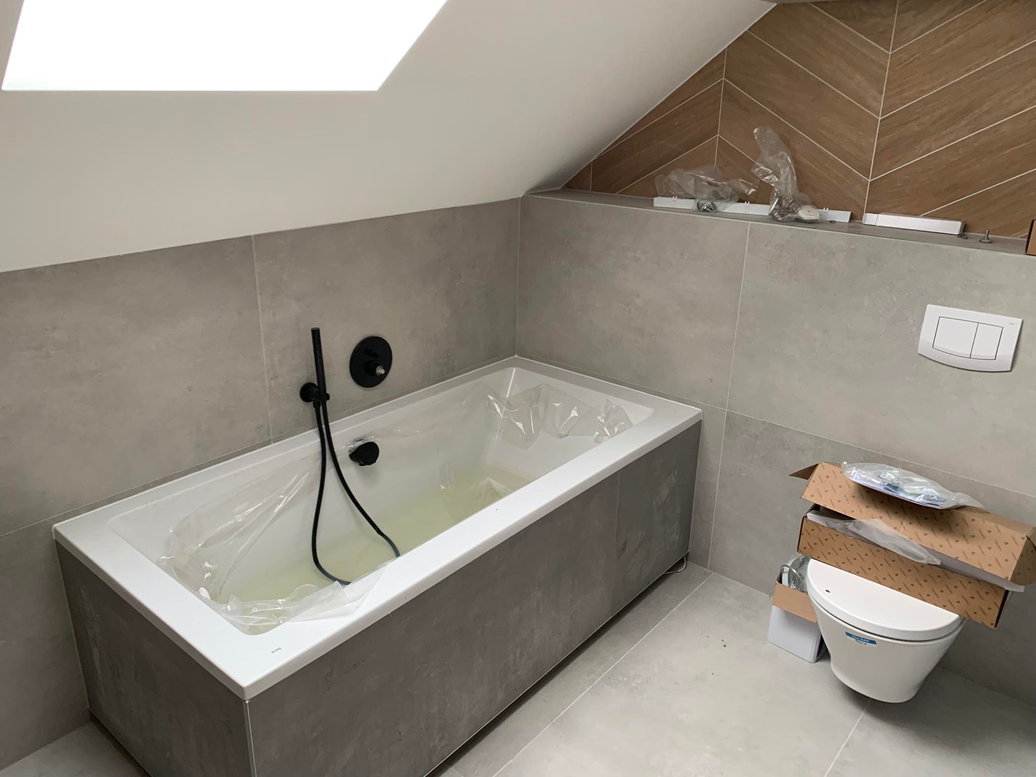 Nowa wanna z czarną armaturą, osłonięta folią ochronną, w łazience z szarymi płytkami i skośnym sufitem z oknem dachowym, obok zapakowana toaleta.
