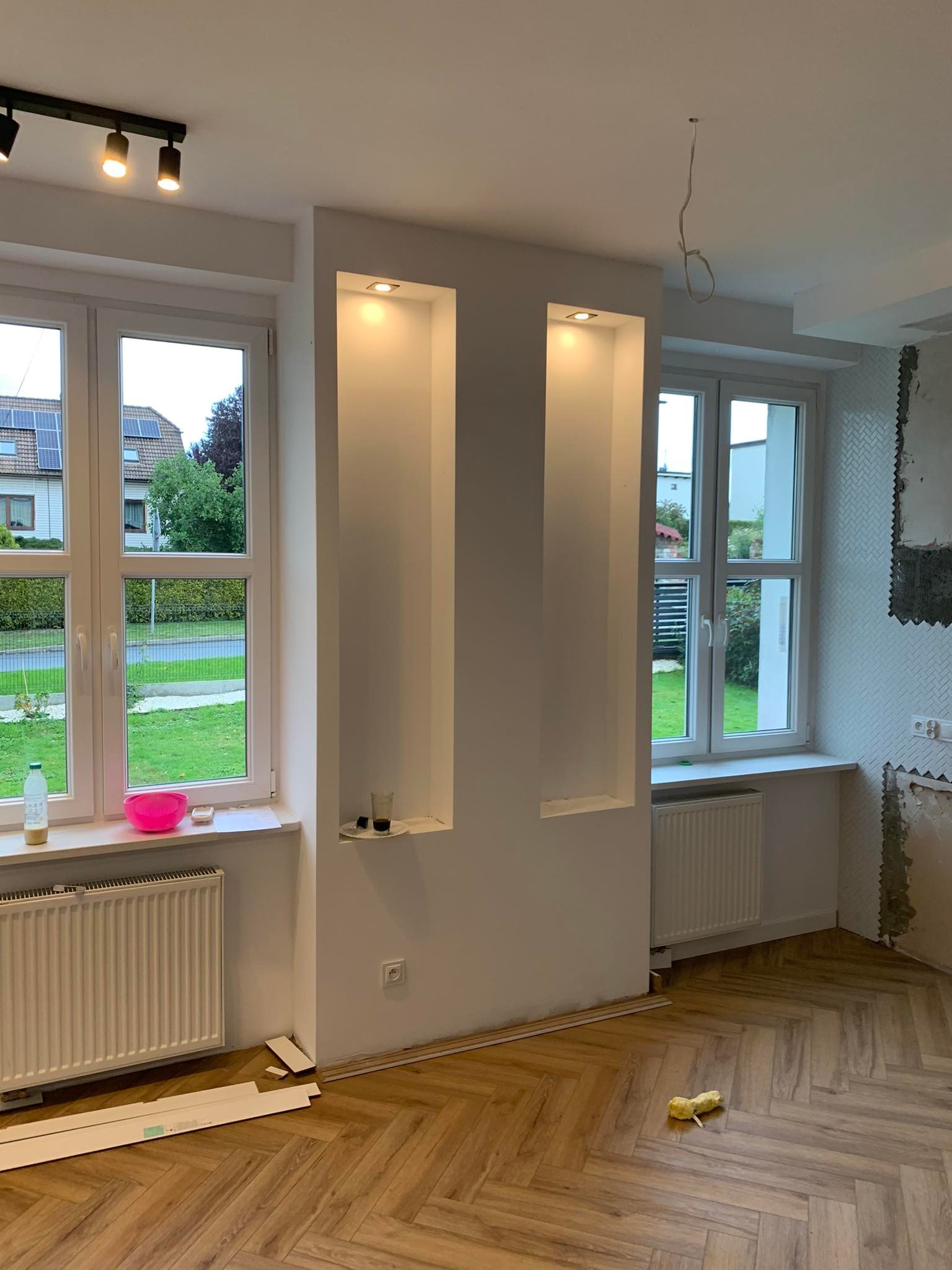 Pokój w trakcie remontu: ułożona podłoga z paneli w jodełkę, wnęki ścienne z oświetleniem, okna z widokiem na ogród, niedokończona ściana z mozaiką, widoczne materiały i narzędzia.