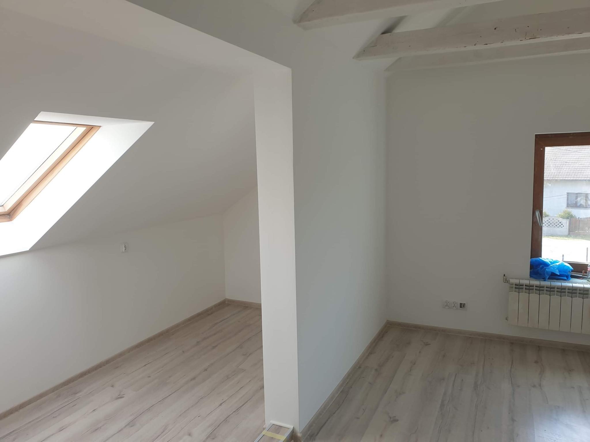 Poddasze po remoncie: świeżo pomalowane białe ściany, drewniane belki stropowe, okno dachowe, grzejnik pod oknem z niebieską folią ochronną, panele podłogowe imitujące drewno.