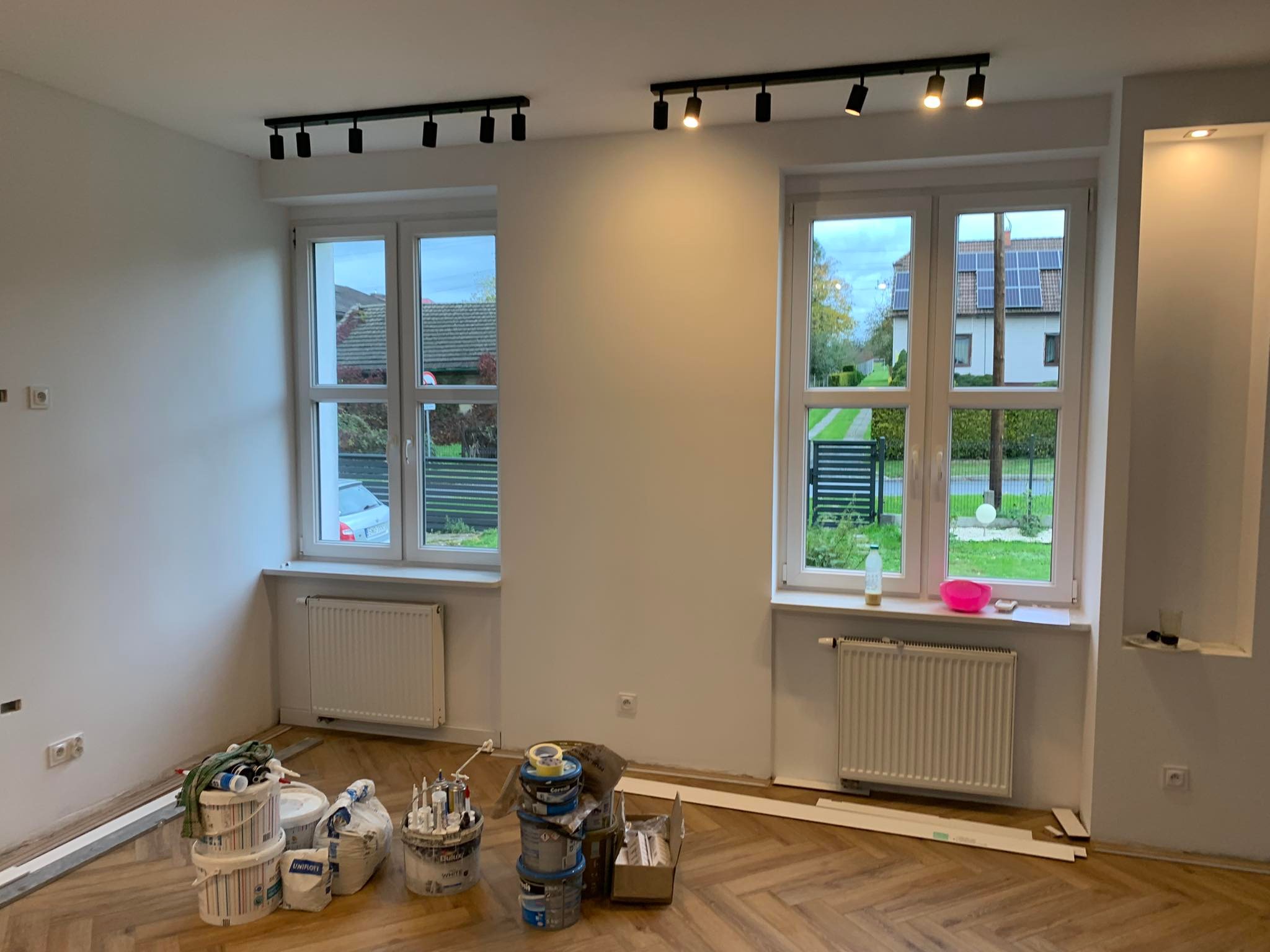 Pomalowane na biało wnętrze pokoju z parkietem w jodełkę, widoczne dwa okna z grzejnikami, puszki z farbą i narzędzia malarskie przy ścianie, oświetlenie szynowe na suficie.