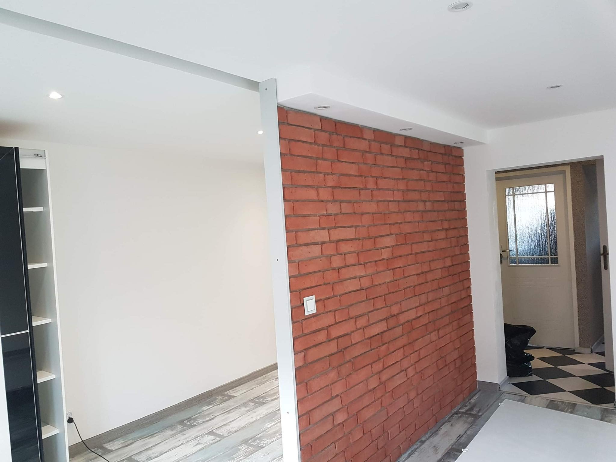 Ściana z cegły dekoracyjnej w odnowionym wnętrzu, widoczne fragmenty podłogi z paneli i drzwi wejściowe.