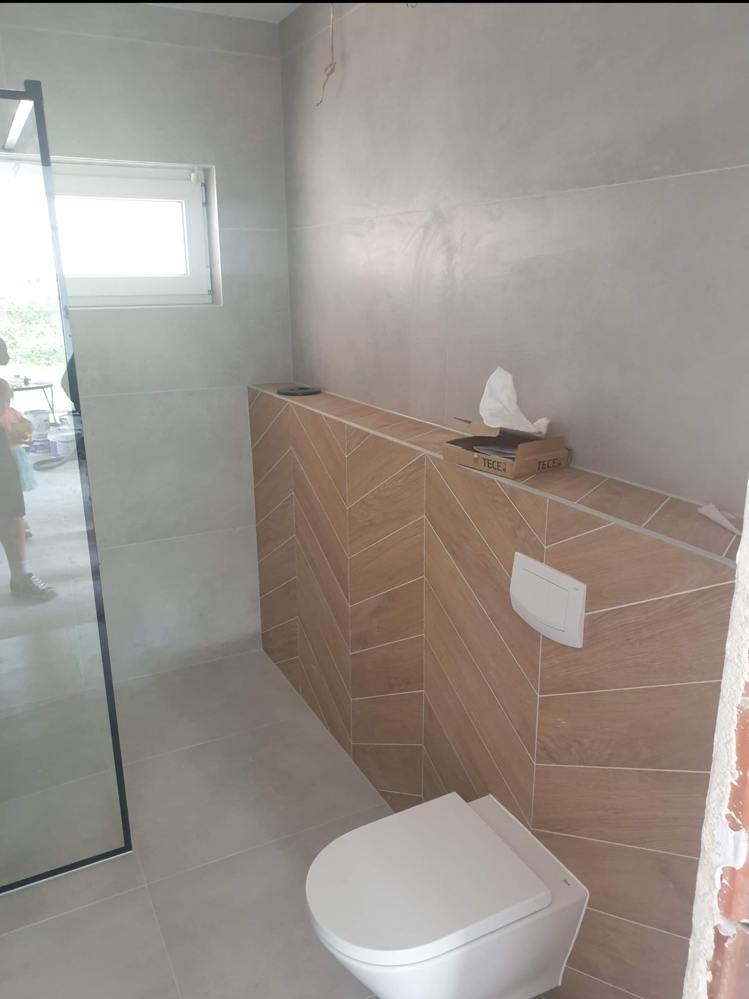 Nowoczesna łazienka w trakcie wykańczania: podwieszana toaleta, drewnopodobne płytki ułożone w jodełkę na zabudowie stelaża, szklana kabina prysznicowa z czarnymi profilami, szare płytki...