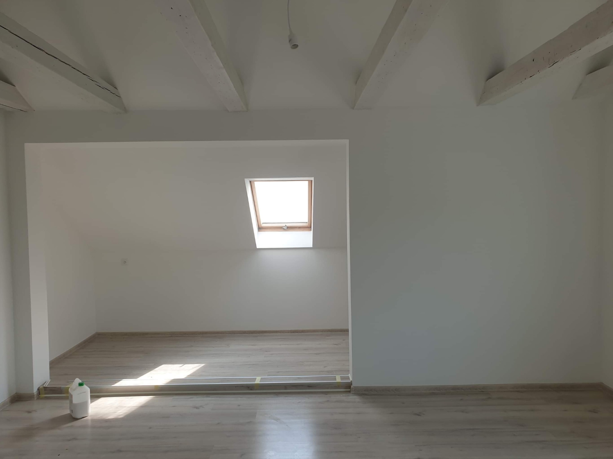 Poddasze po remoncie z drewnianymi belkami stropowymi, jasnymi panelami podłogowymi i oknem dachowym, widoczna poziomica i pojemnik z farbą.