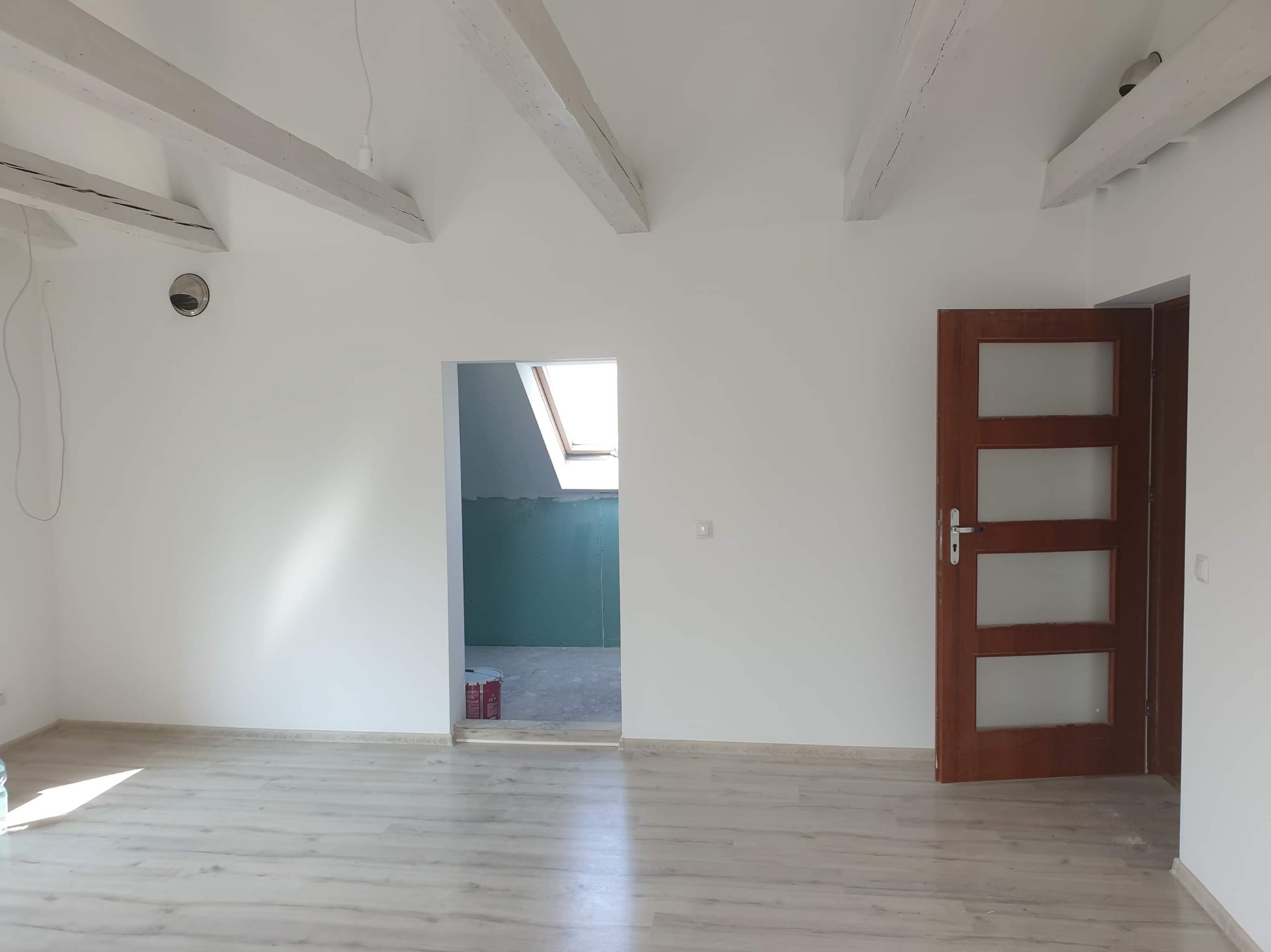 Poddasze po remoncie: białe ściany, drewniane belki stropowe, jasna podłoga z paneli, widoczne drzwi wewnętrzne z przeszkleniami i fragment pomieszczenia z oknem dachowym i niepomalowanymi ścianami.