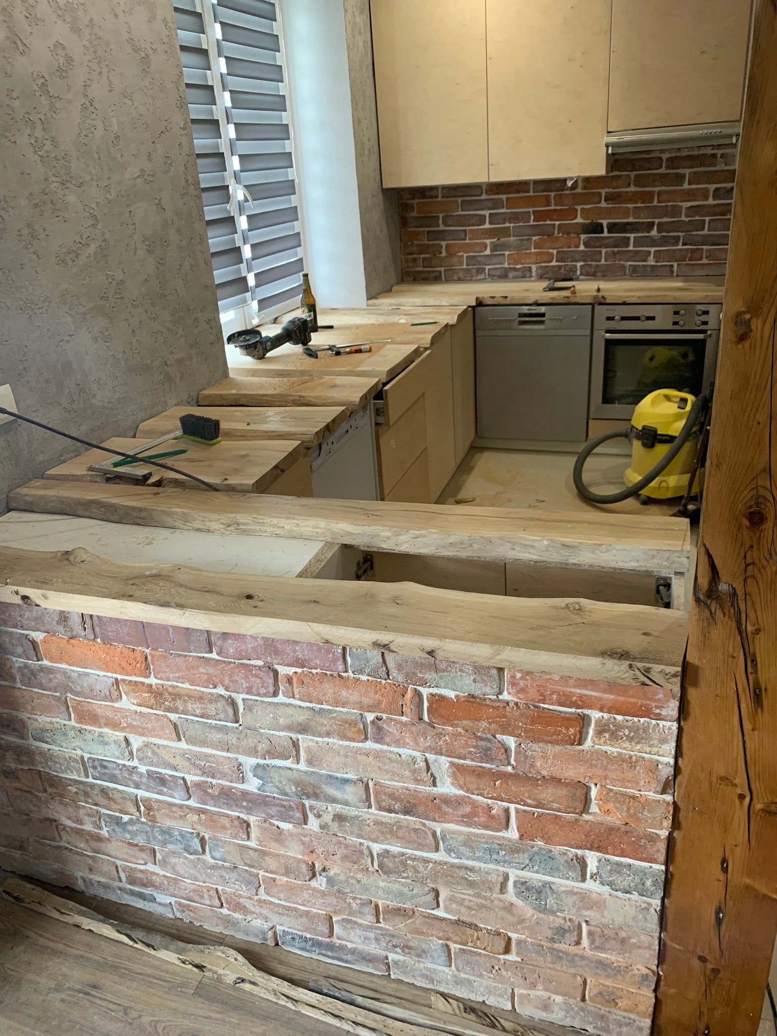 Wnętrze kuchni w trakcie montażu drewnianego blatu z surowego drewna, widoczny mur z cegły, narzędzia, butelka oraz odkurzacz przemysłowy.