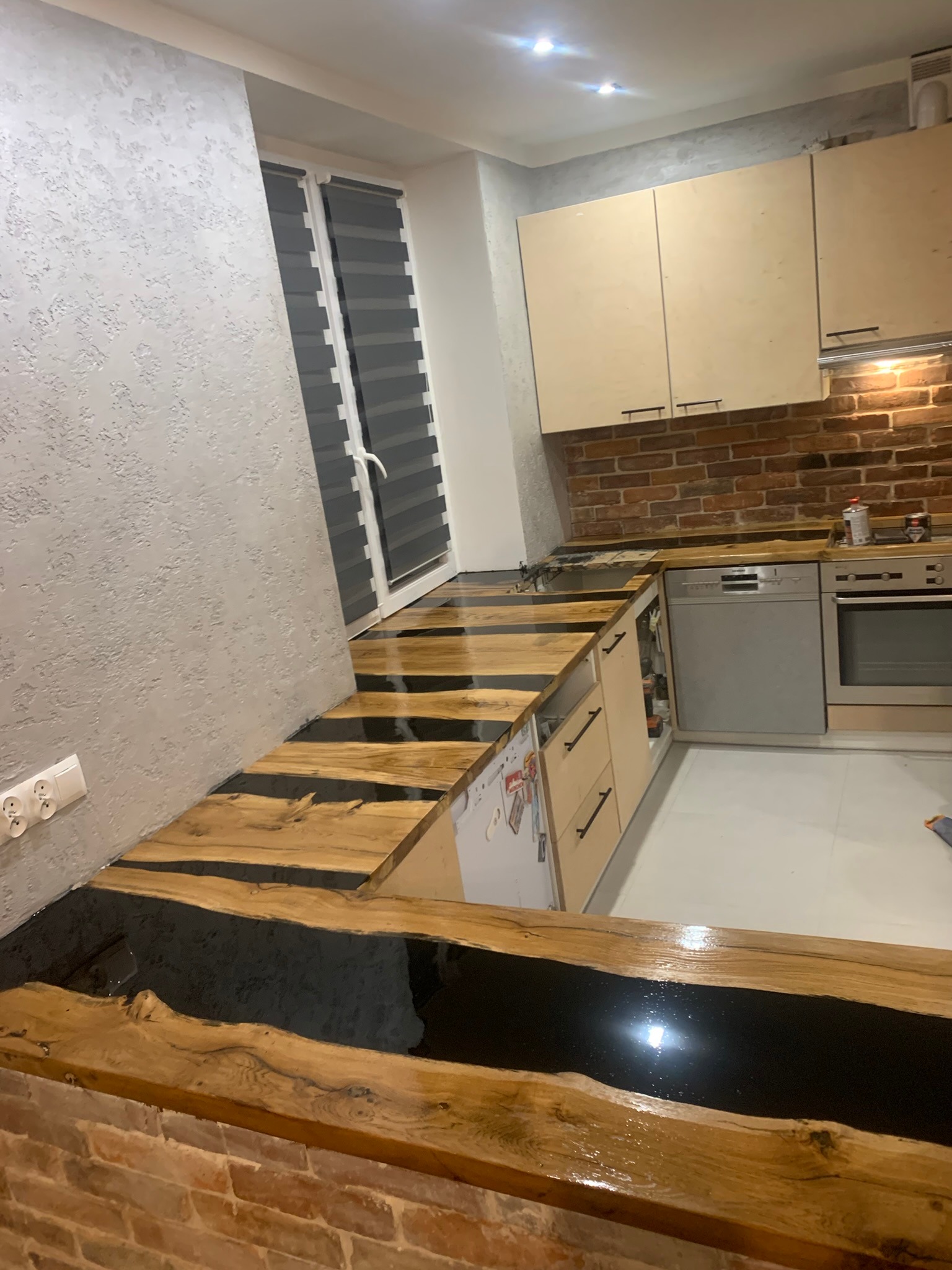 Nowoczesna kuchnia z blatem z drewna i żywicy epoksydowej, ściana z cegły, jasne szafki i szara ściana z fakturą.