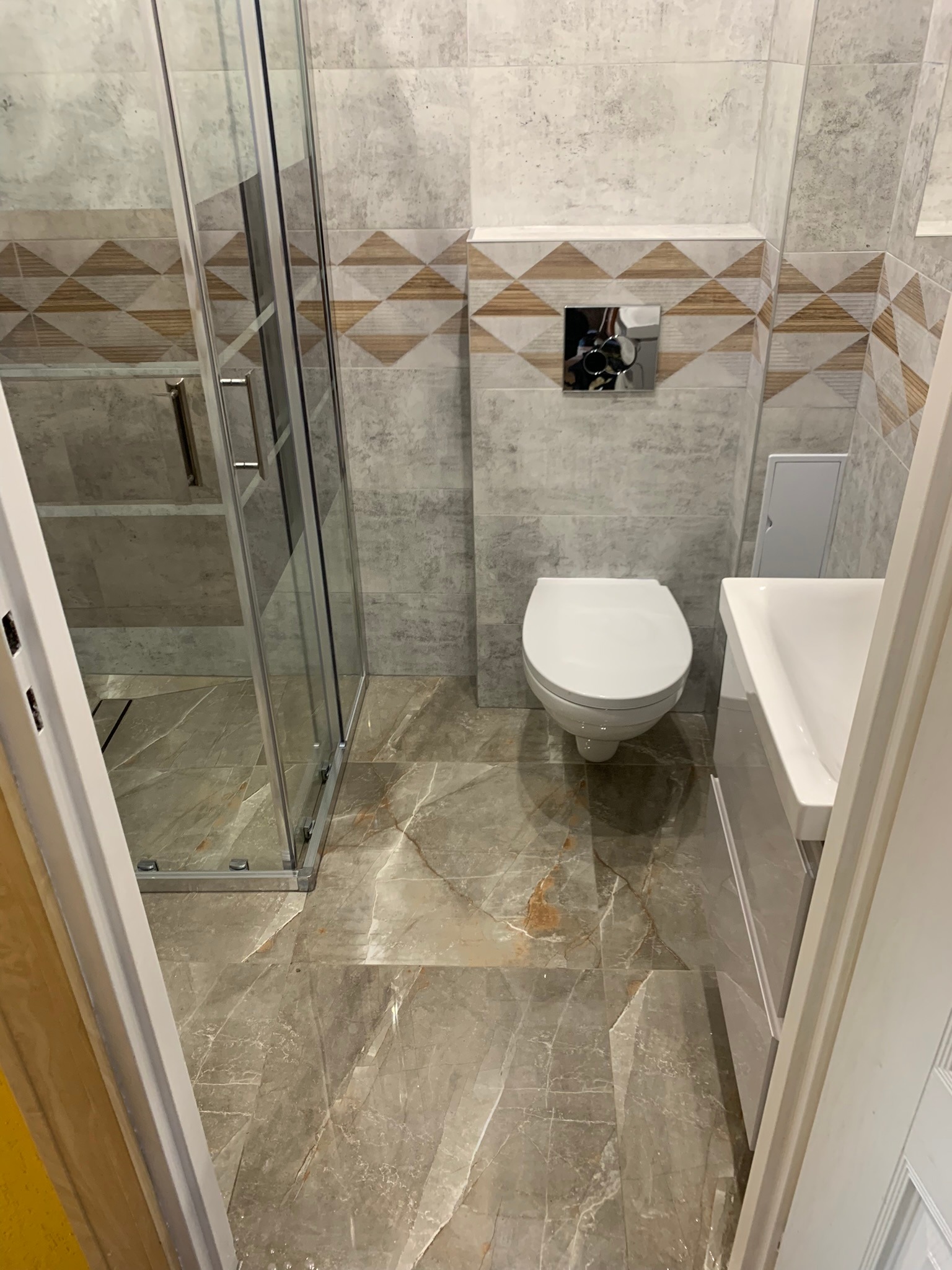 Nowoczesna łazienka z kabiną prysznicową, wiszącą toaletą i umywalką w zabudowie, wykończona płytkami imitującymi marmur z dekoracyjnym pasem z geometrycznym wzorem.