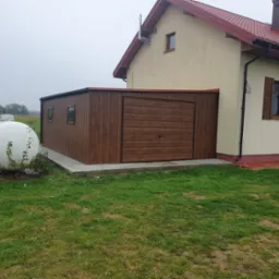 Garaż blaszany z bramą segmentową w kolorze drewna, przylegający do ściany domu, z widocznym zbiornikiem na gaz w tle.