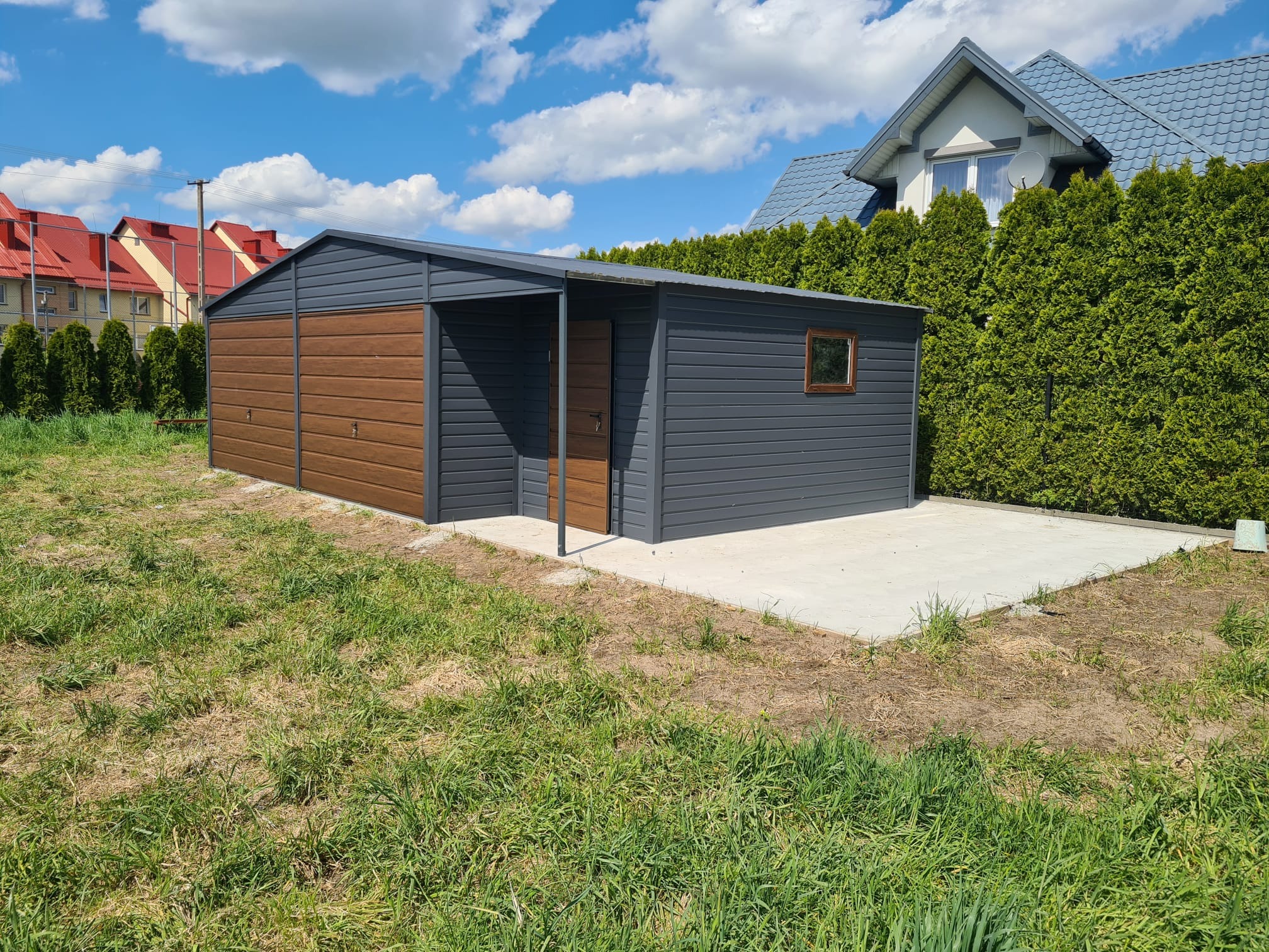 Szary garaż blaszany z drewnopodobnymi drzwiami segmentowymi i wejściowymi, z oknem, na betonowej wylewce, otoczony trawnikiem, w tle dom z szarym dachem i zielone tuje.