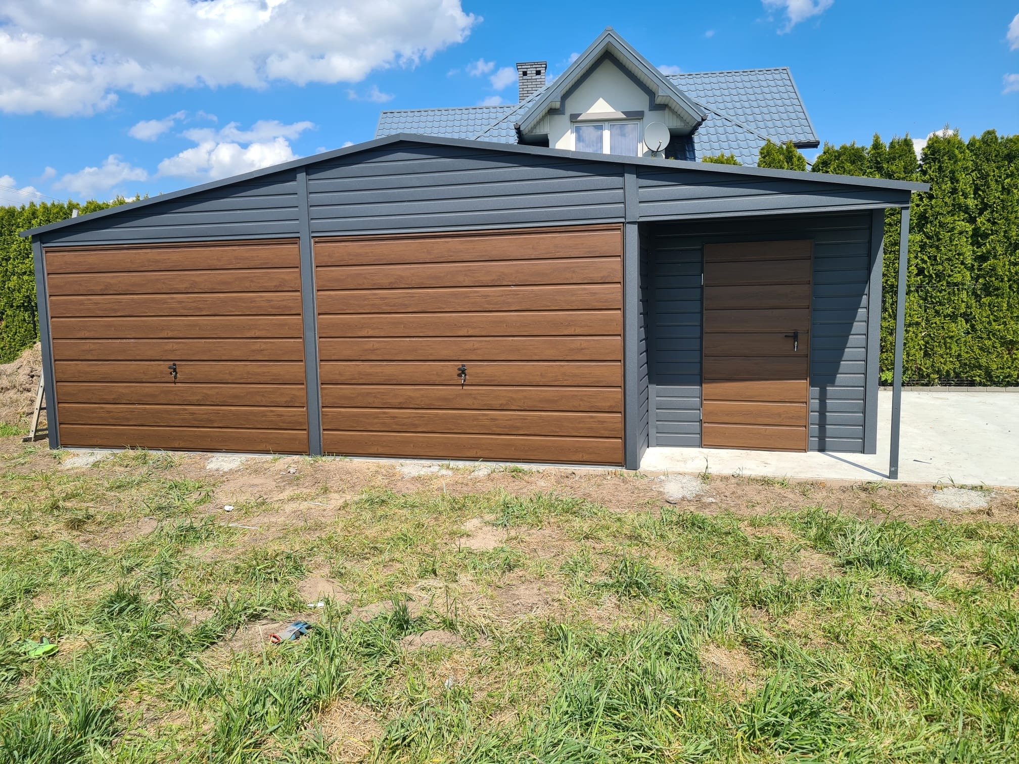 Dwustanowiskowy garaż z brązowymi drzwiami segmentowymi i szarą konstrukcją, zlokalizowany w Raciechowicach, na tle domu jednorodzinnego z szarym dachem i zielonymi tujami.