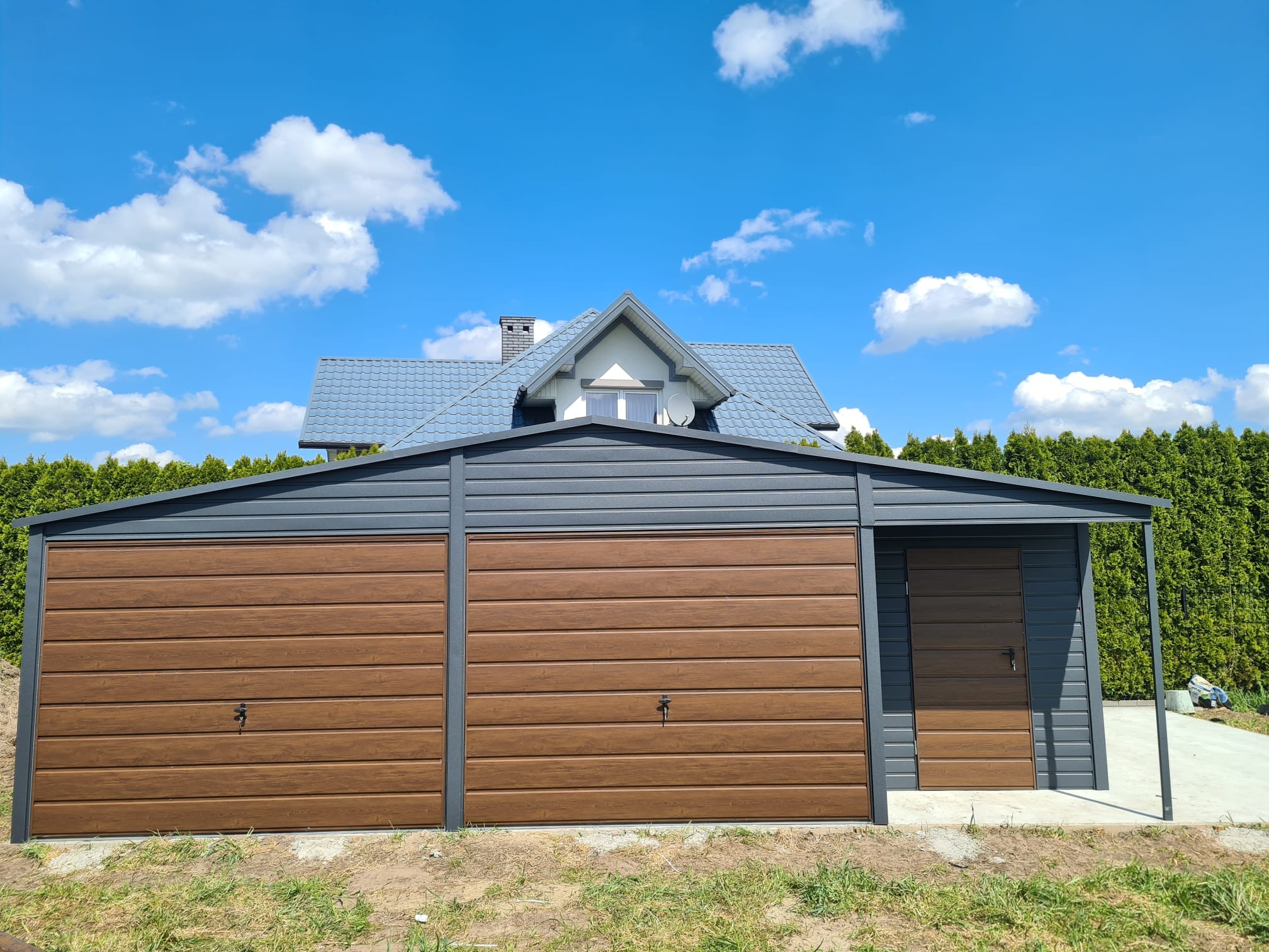 Nowoczesny garaż blaszany z dwoma bramami segmentowymi w kolorze drewna, z dodatkowym wejściem bocznym, na tle domu i zielonych tuj.
