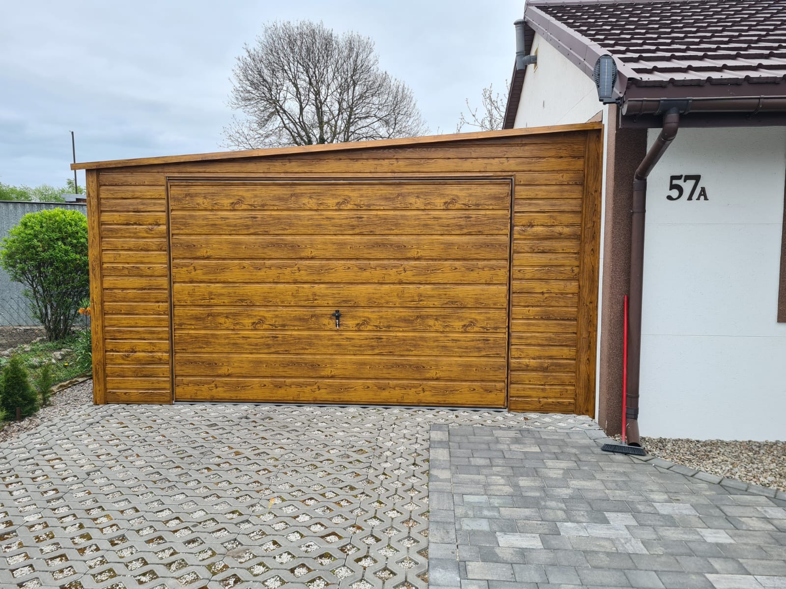 Gotowy garaż blaszany z imitacją drewna, zamek na środku drzwi, kostka ażurowa przed garażem, obok budynku z numerem 57A.