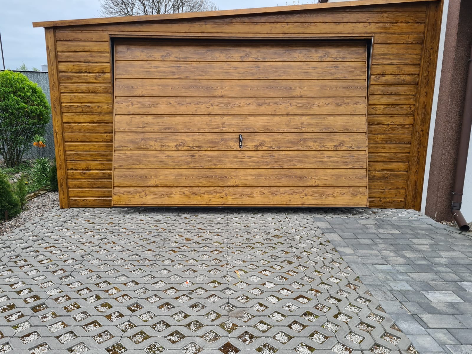 Gotowy garaż z blachy imitującej drewno, zamykany na pojedynczy zamek, położony na nawierzchni z ażurowych płyt wypełnionych drobnym kamieniem.