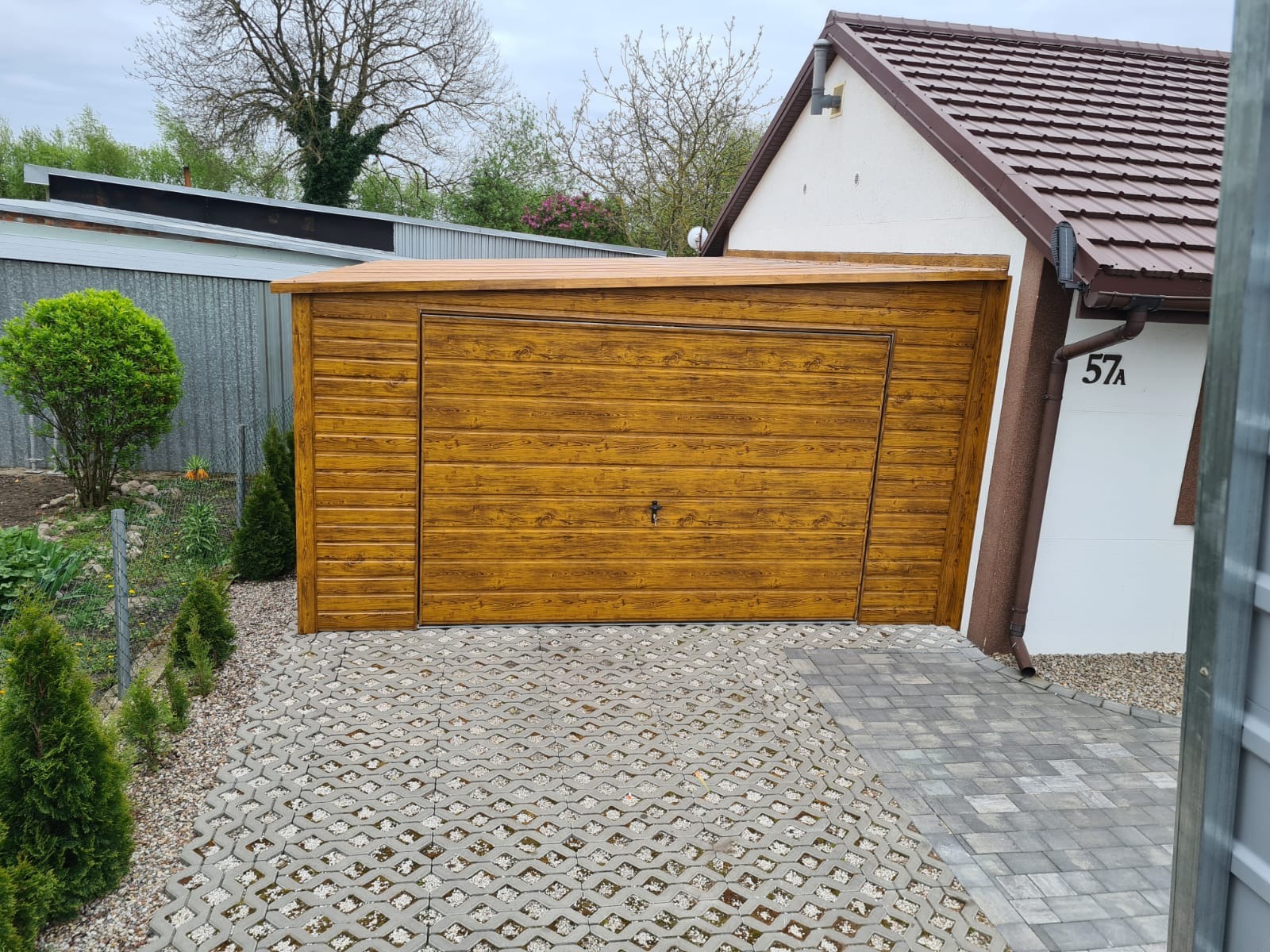 Drewnopodobny garaż z poziomymi panelami, zamykany na klucz, umieszczony na ażurowej kostce brukowej obok budynku z numerem 57A.