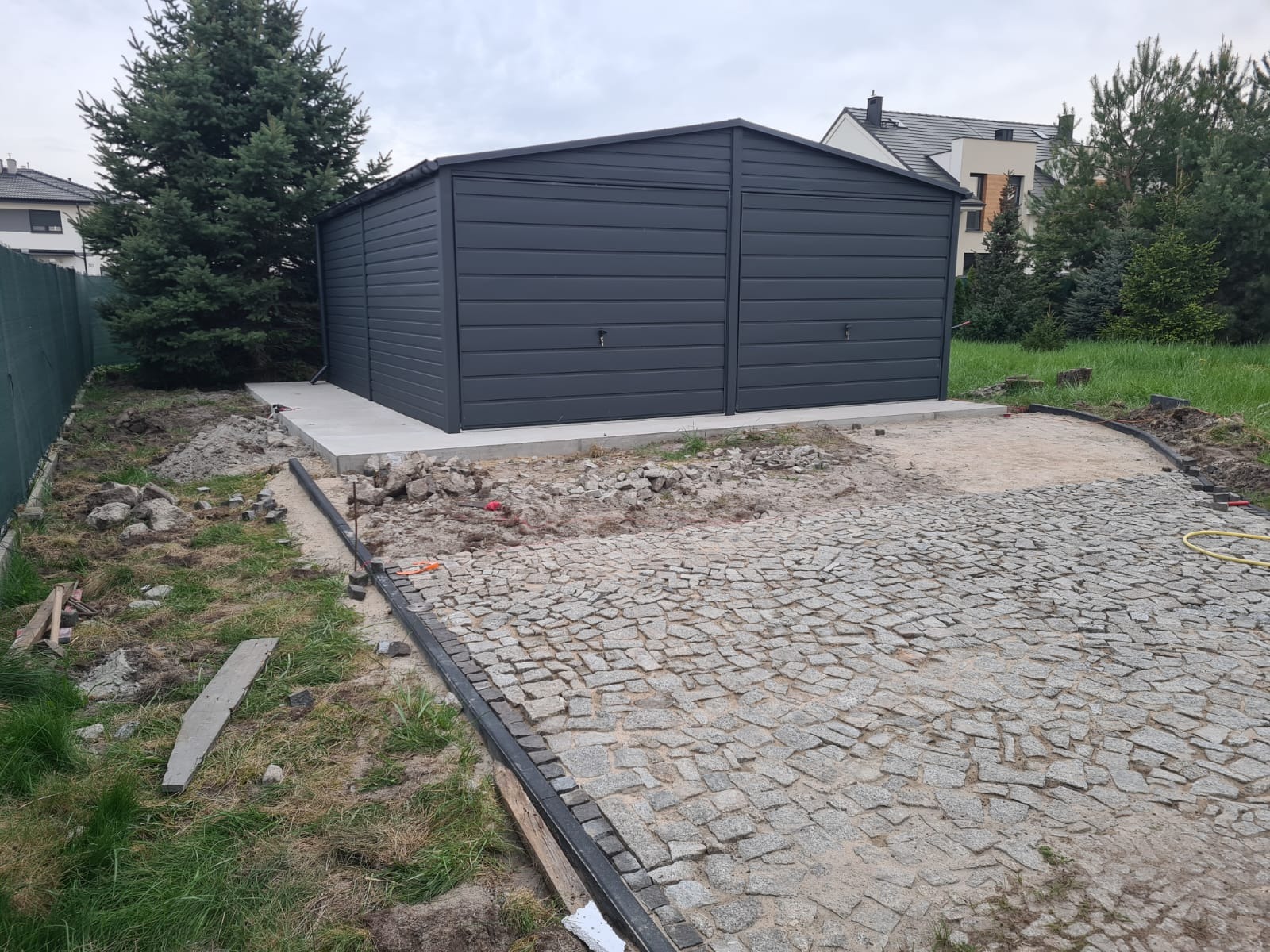 Szary garaż z blachy falistej z dwoma bramami, ustawiony na betonowej wylewce, widok na posesję z kostką brukową w trakcie układania, obok rosną drzewa iglaste.