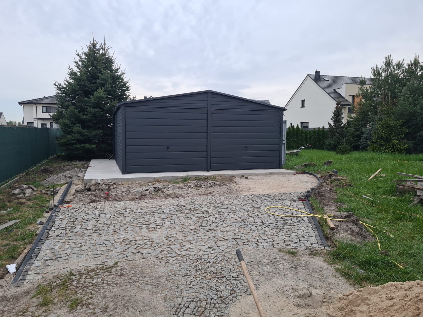 Dwustanowiskowy garaż blaszany w kolorze grafitowym na betonowej wylewce, widoczny w trakcie budowy podjazdu z kostki granitowej, w tle dom jednorodzinny i iglak.