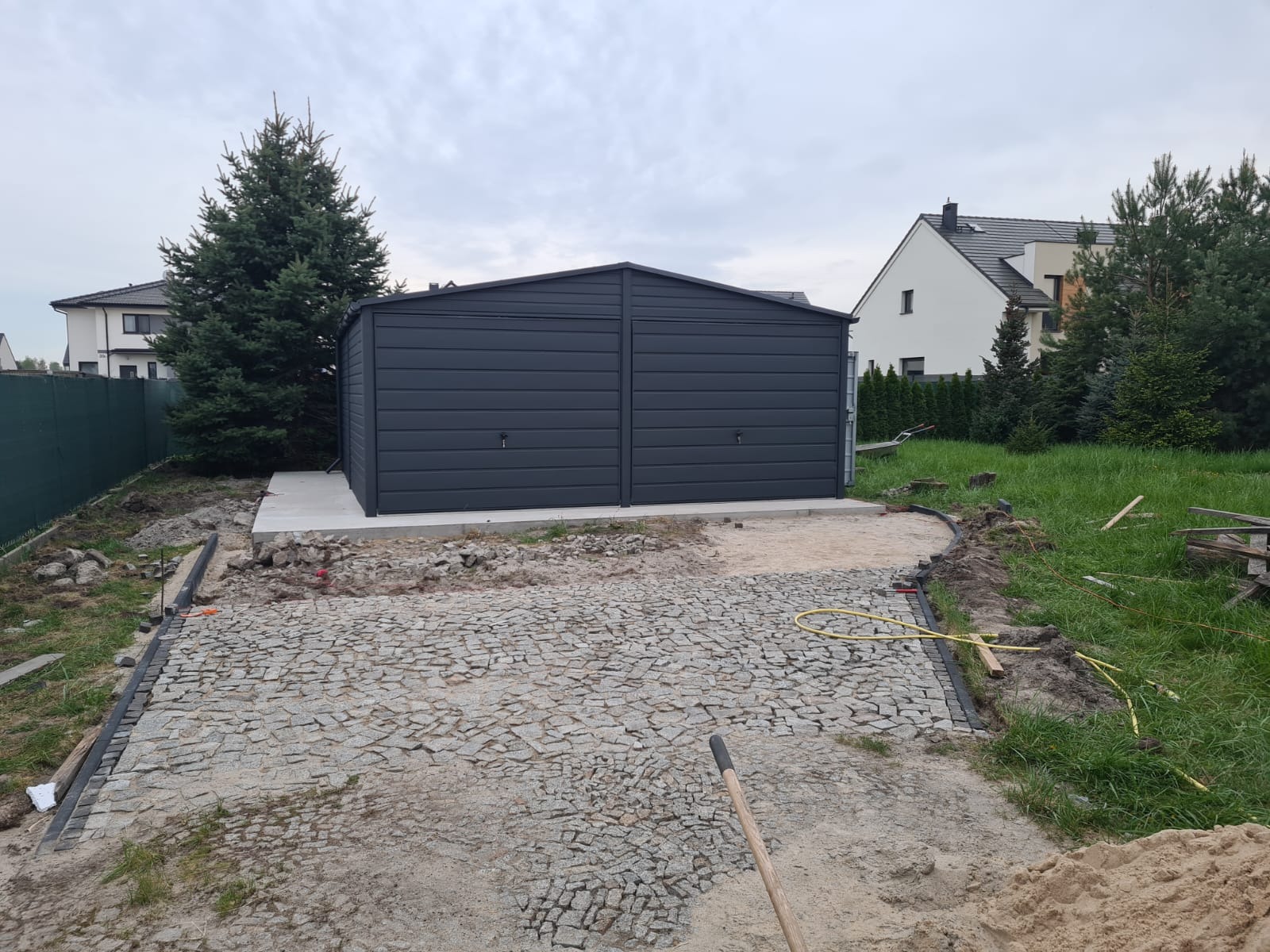 Dwustanowiskowy, grafitowy garaż blaszany na betonowej wylewce, widok z poziomu ziemi podczas układania kostki brukowej na podjeździe.