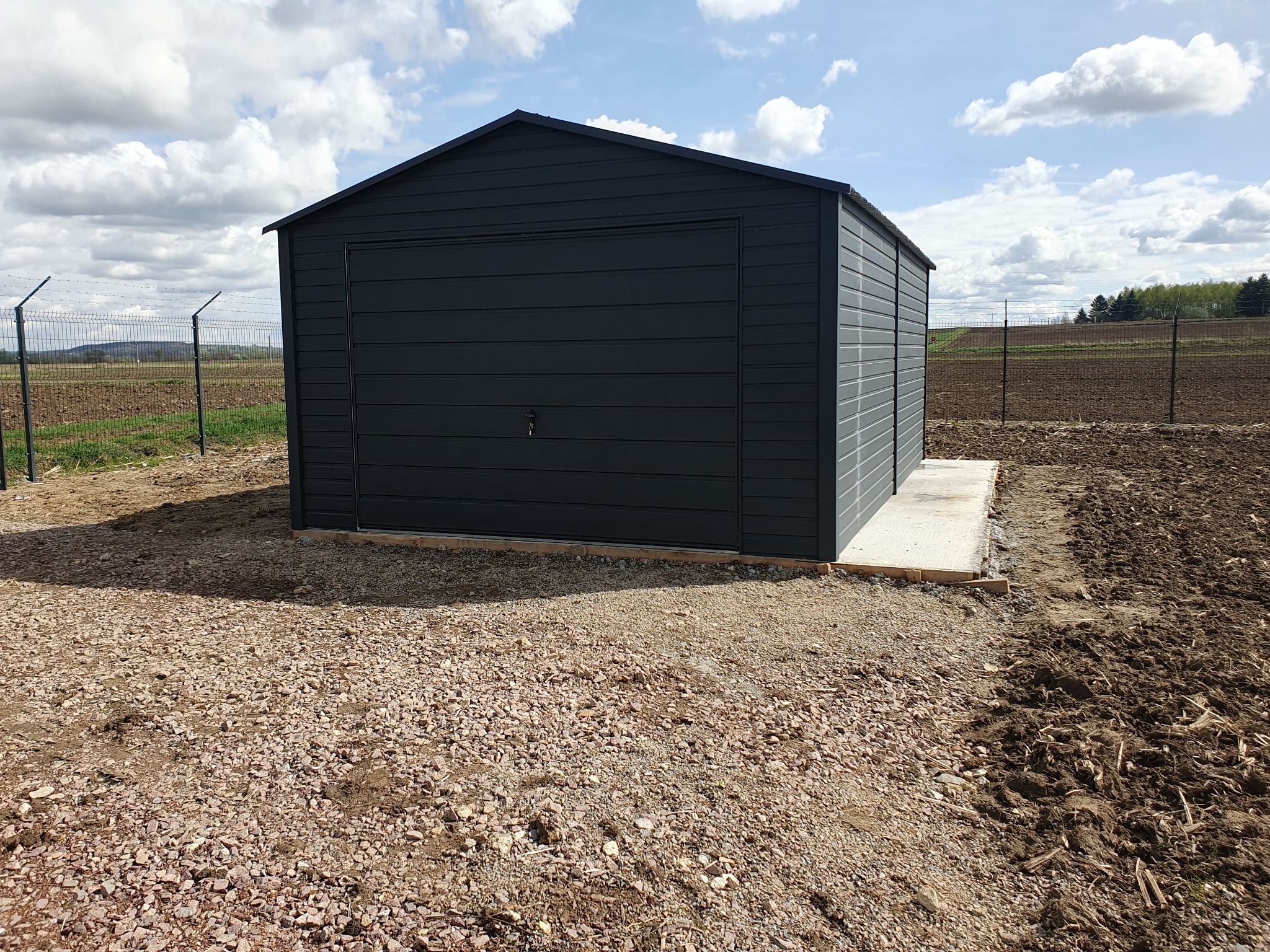 Nowoczesny, grafitowy garaż blaszany z pojedynczą bramą, umieszczony na betonowej wylewce na żwirowym podłożu, z widocznym ogrodzeniem z siatki w tle i pochmurnym niebem.