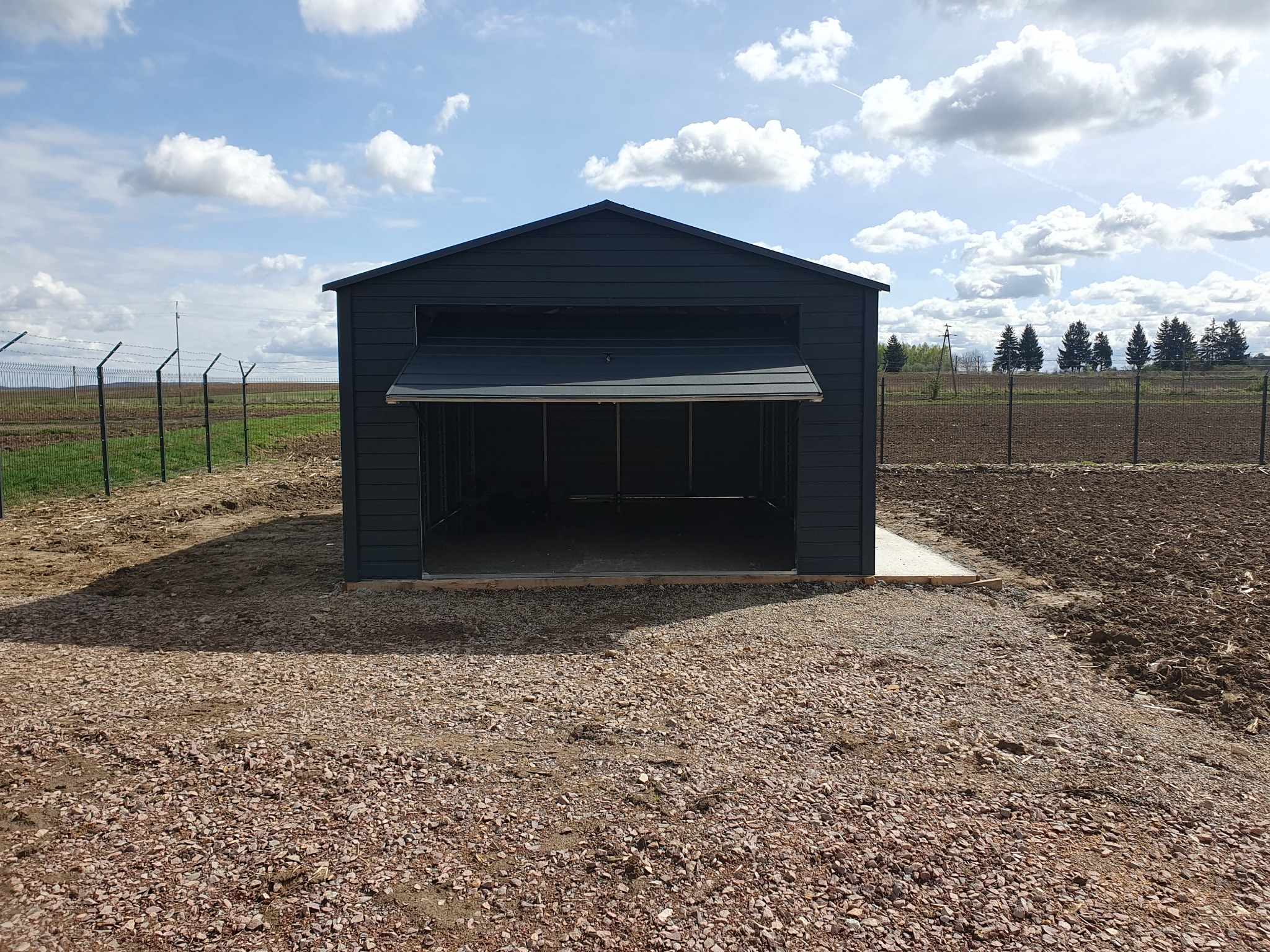 Nowoczesny, antracytowy garaż z blachy trapezowej z uchylną bramą, ustawiony na utwardzonym żwirowym podłożu, w tle pole i ogrodzenie z siatki.