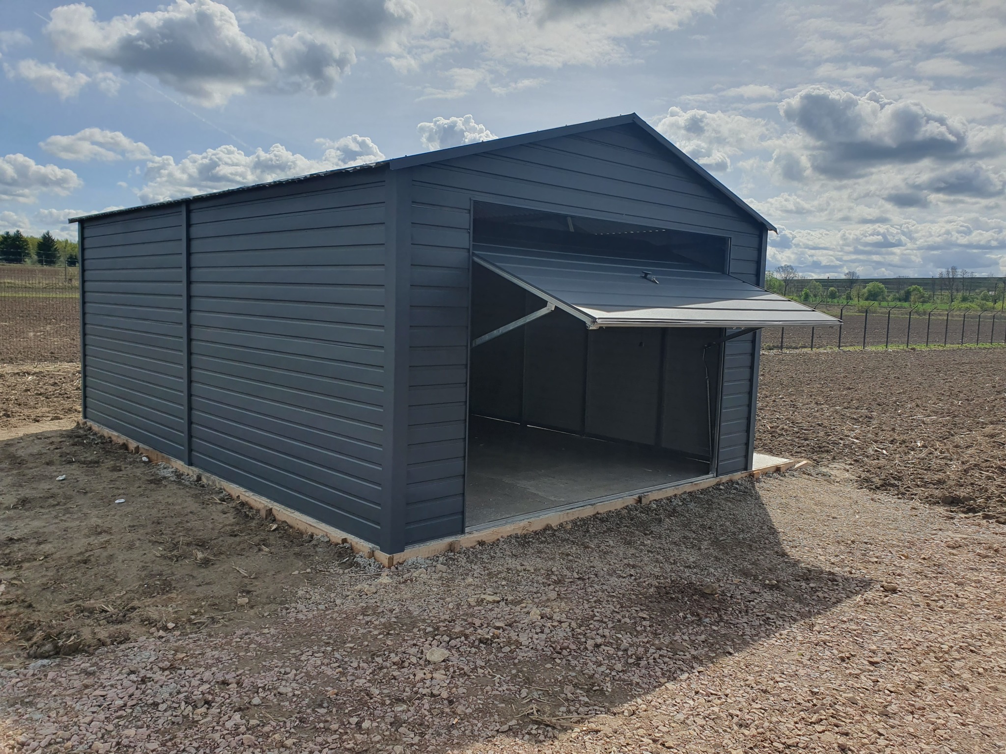 Nowoczesny, antracytowy garaż blaszany z uchyloną bramą, ustawiony na betonowej wylewce na tle zaoranego pola i błękitnego nieba z chmurami.