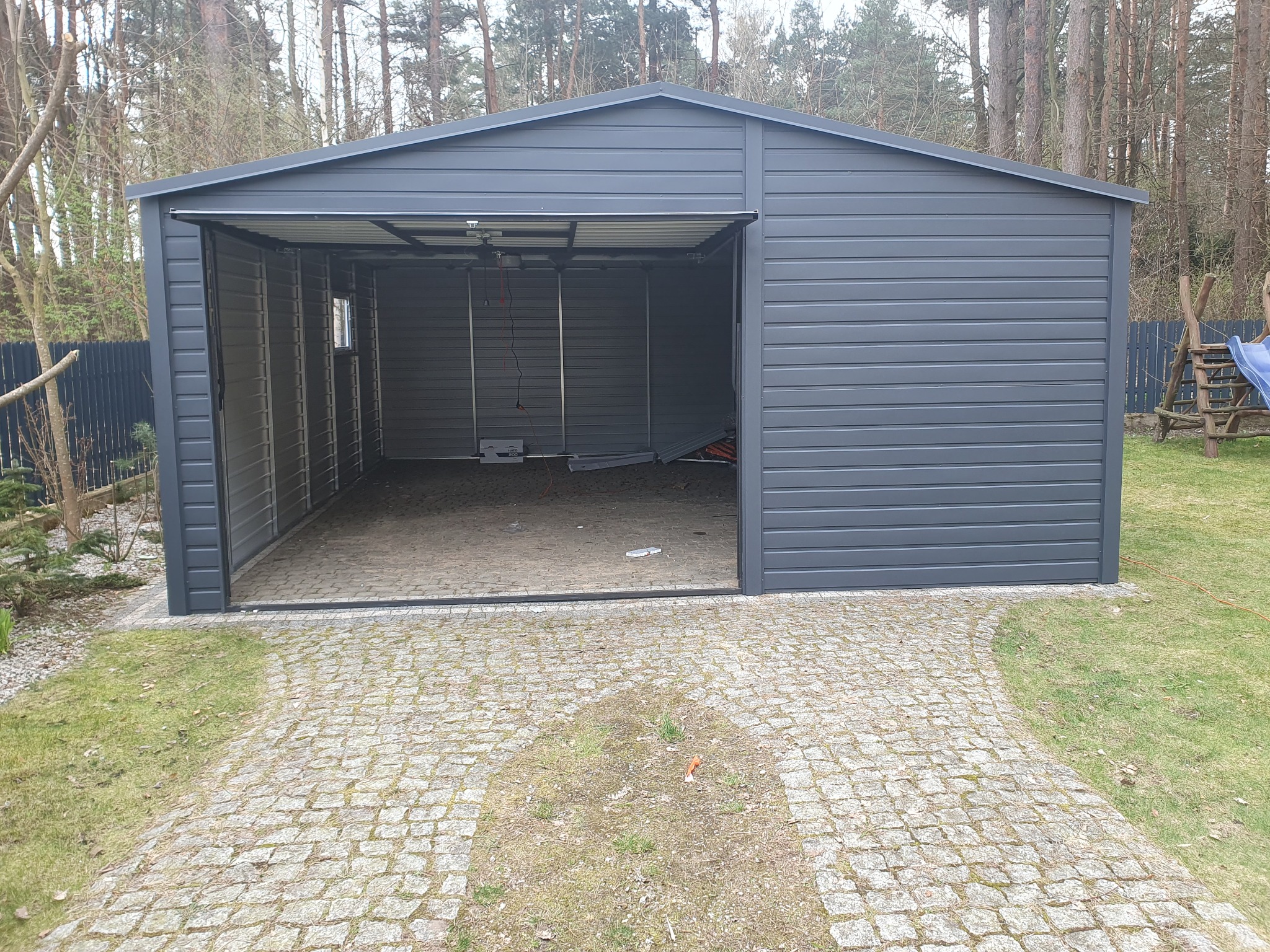 Szary garaż z blachy falistej, otwarty, z widocznym wnętrzem i brukowanym podjazdem na posesji z drzewami w tle.