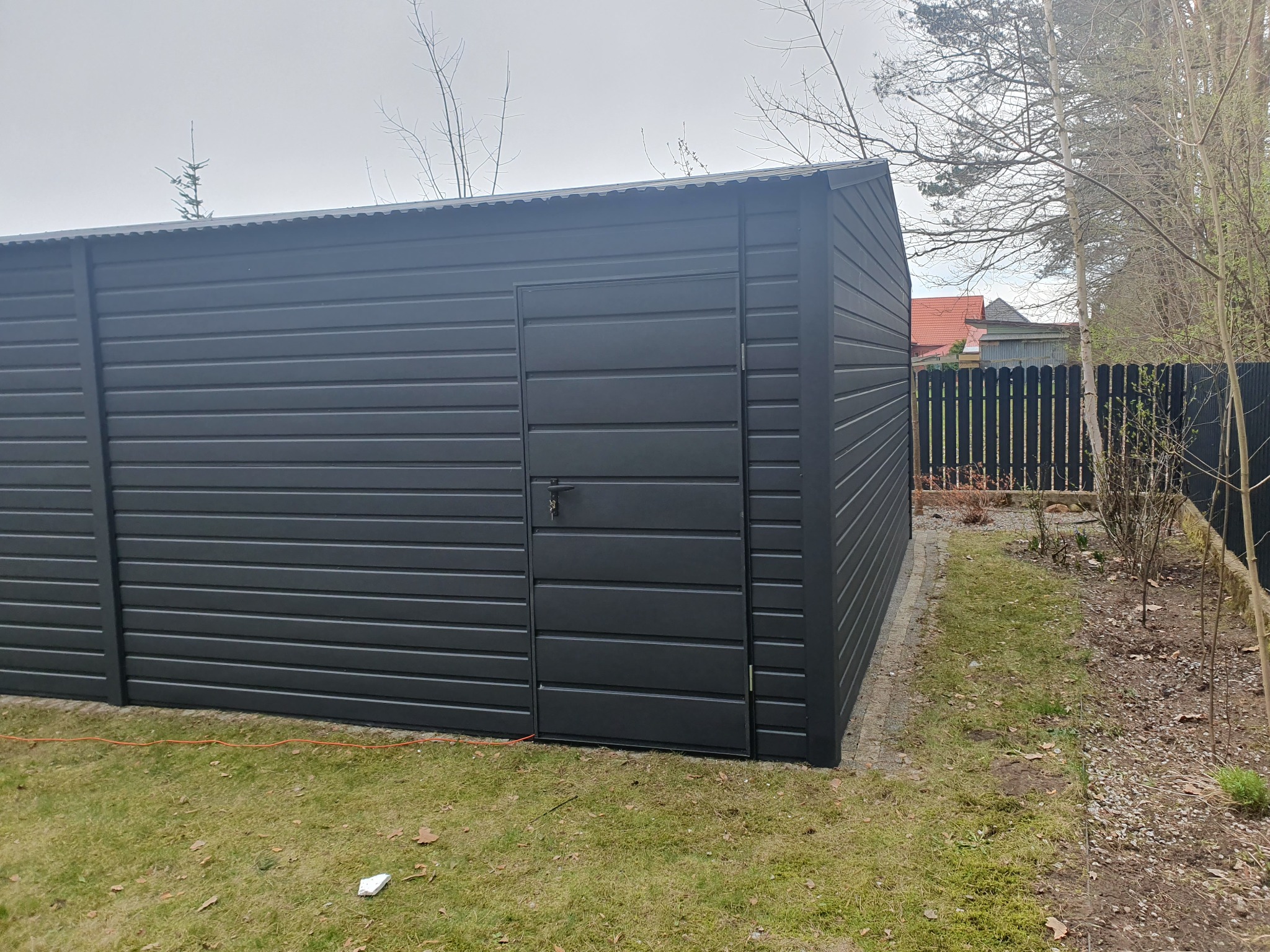Nowoczesny, grafitowy garaż z blachy falistej, widziany z boku na tle trawnika i ogrodzenia z desek. Widoczny kabel elektryczny leżący na trawie przy ścianie garażu.