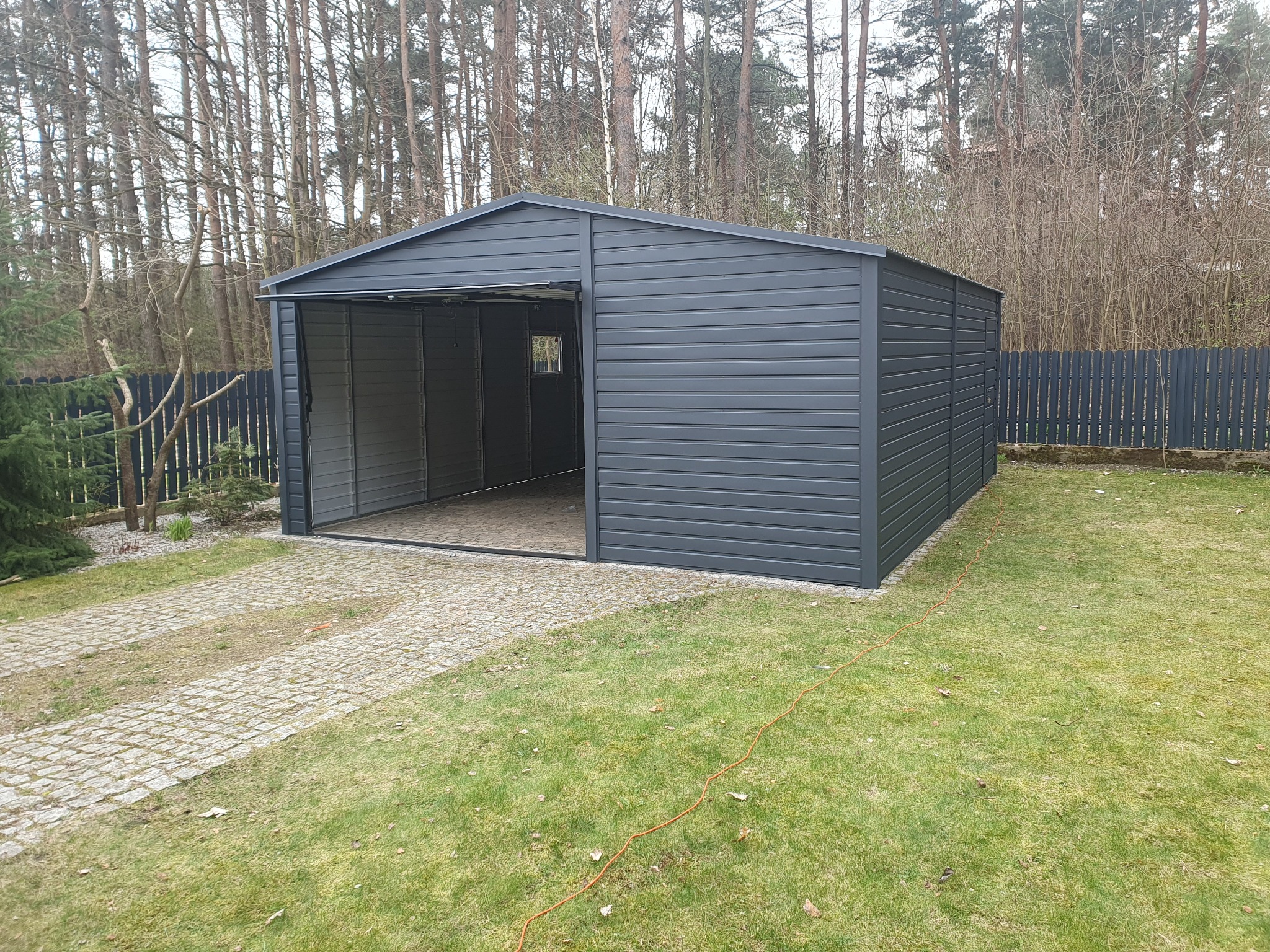 Szary garaż blaszany z otwartymi drzwiami na posesji z kostki brukowej i trawnika, widoczny kabel elektryczny na trawie.
