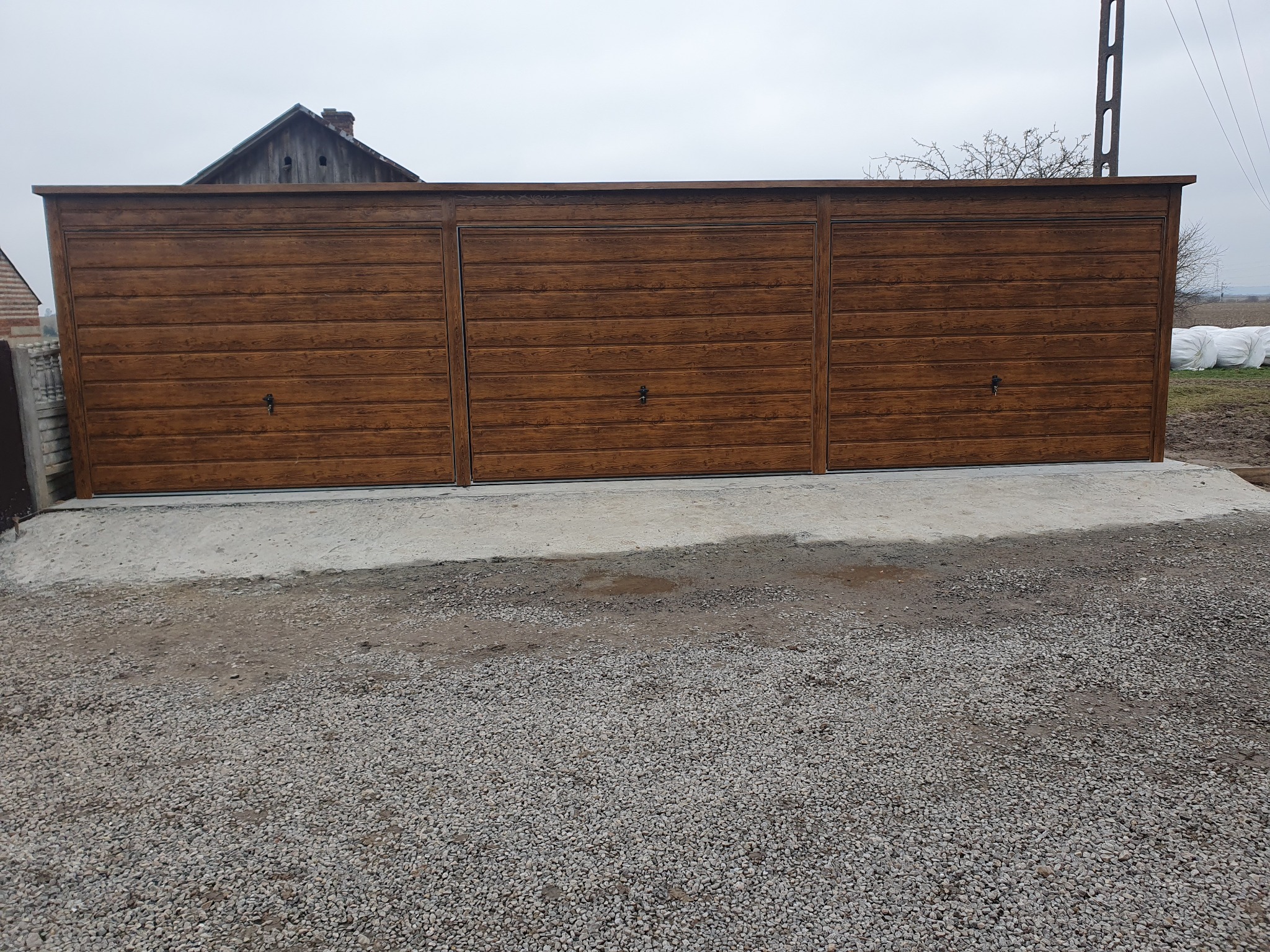 Zewnętrzny, brązowy garaż z blachy imitującej drewno, podzielony na trzy segmenty z uchwytami, posadowiony na betonowej wylewce, z widocznym żwirowym podjazdem i fragmentem domu w tle.