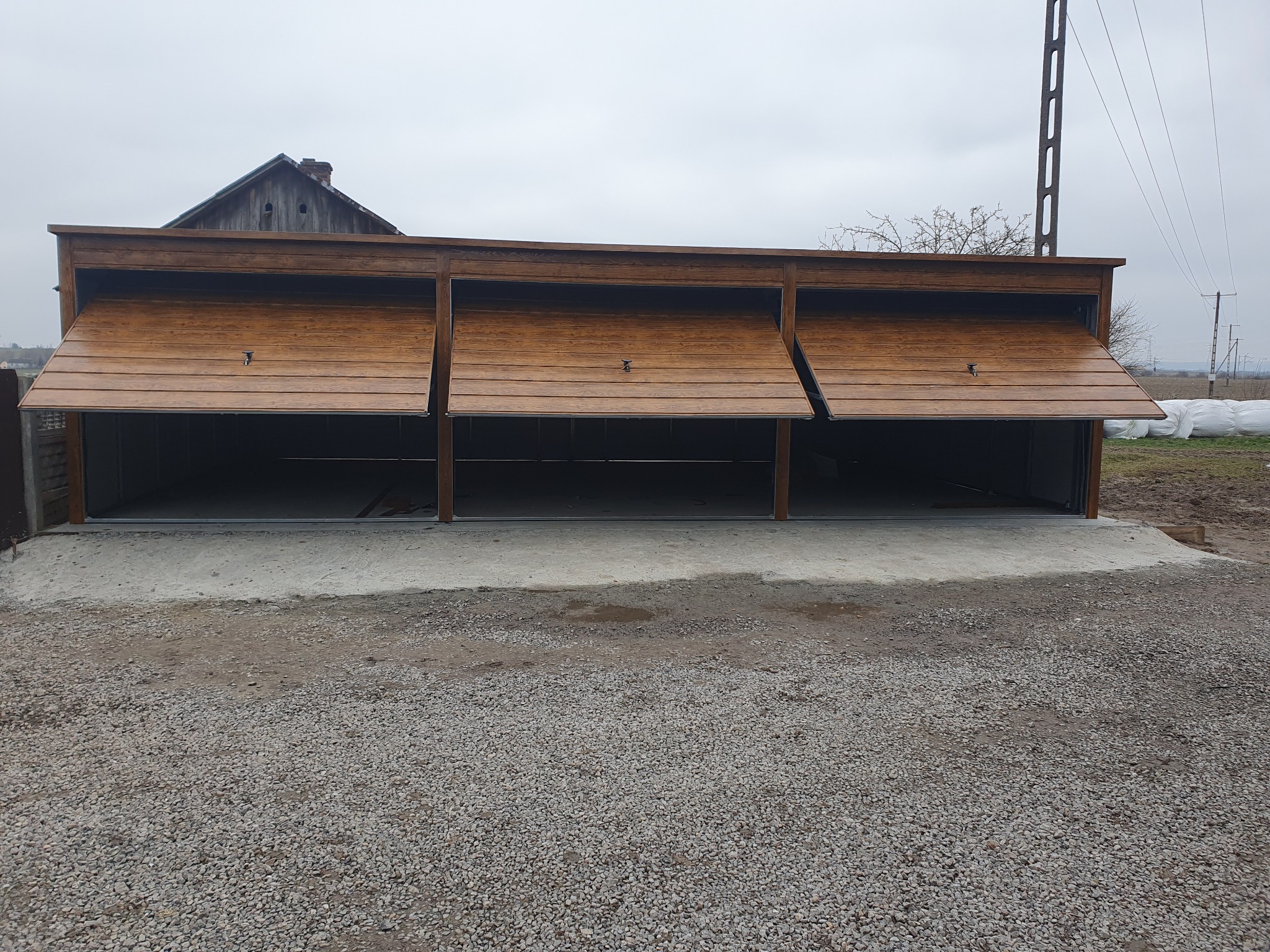 Trzyczęściowy garaż z drewnianymi, częściowo otwartymi drzwiami segmentowymi, widok z zewnątrz na żwirowe podłoże i wiejski krajobraz w tle.