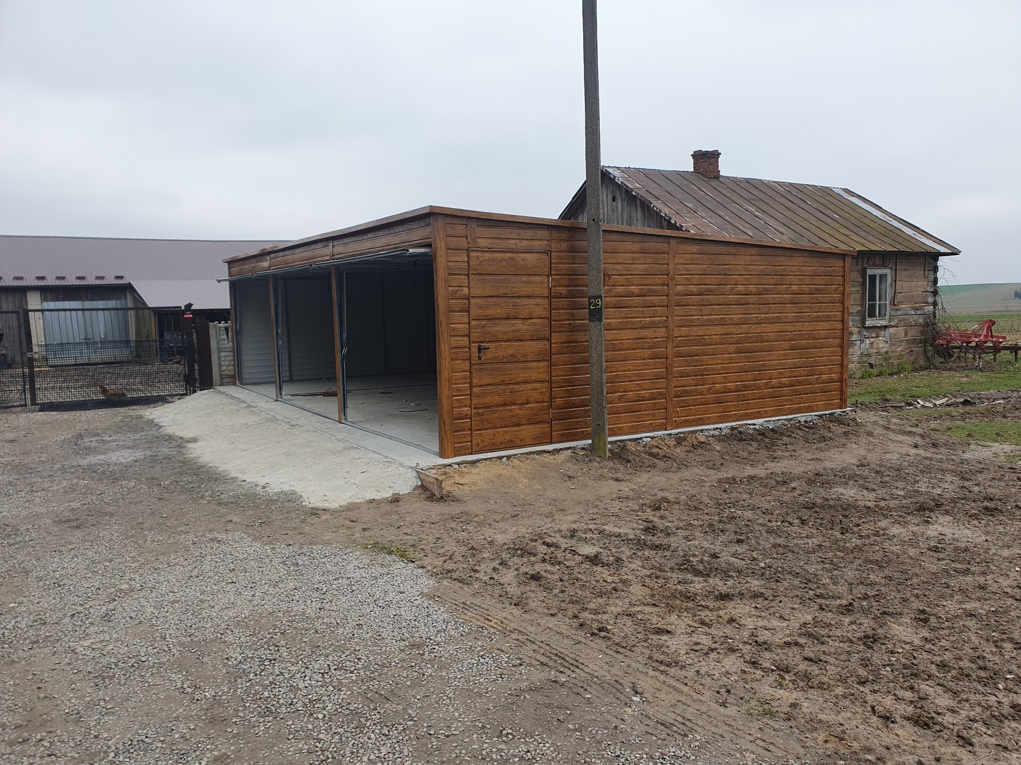 Nowoczesny garaż z drewnopodobną elewacją, częściowo otwarty, z wylewką betonową przed wjazdem, na tle wiejskiego krajobrazu z drewnianym domem i polem.