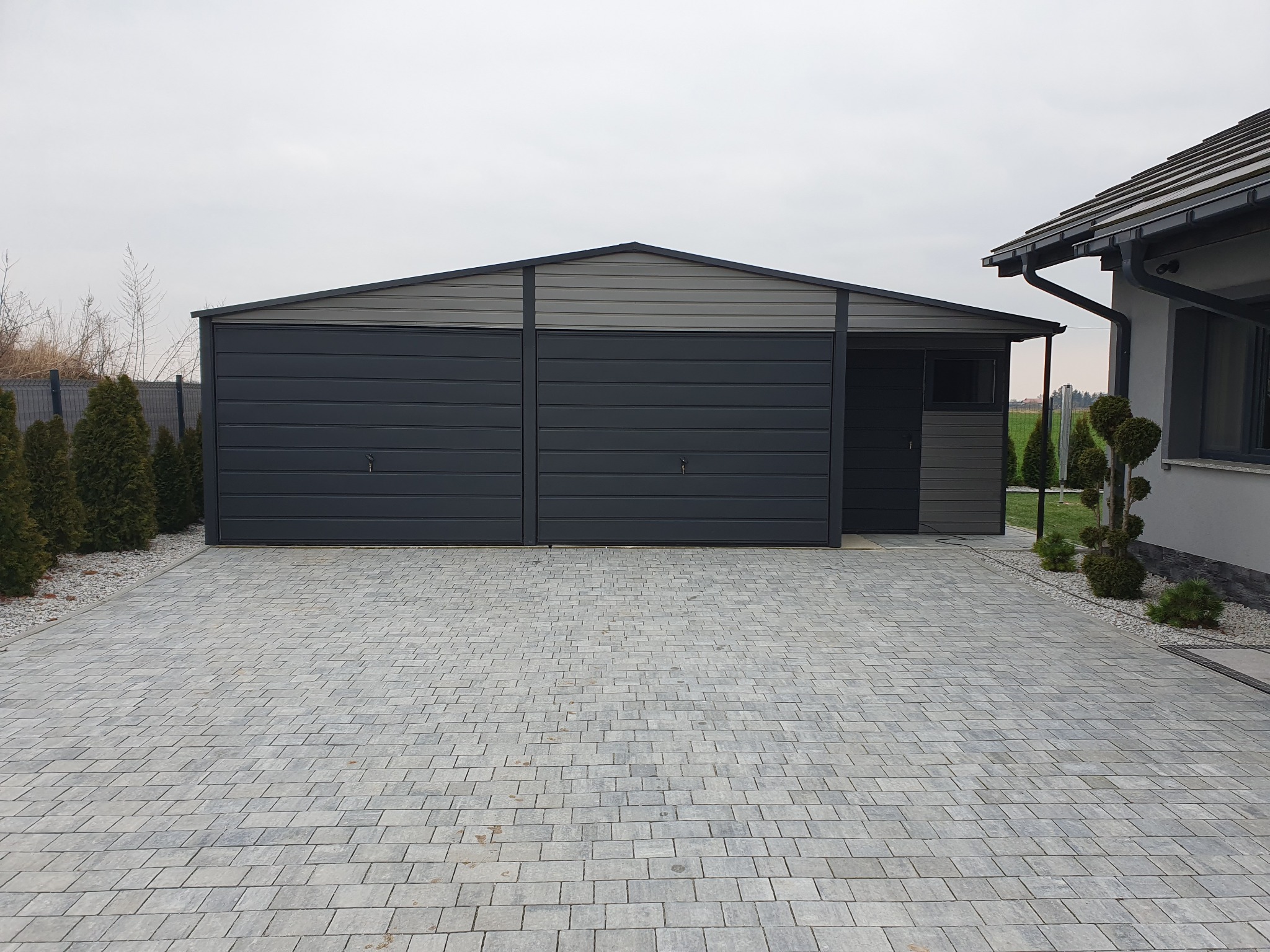 Nowoczesny, dwustanowiskowy garaż z blachy w odcieniach szarości, z dwuspadowym dachem, umieszczony na posesji z wybrukowanym podjazdem.