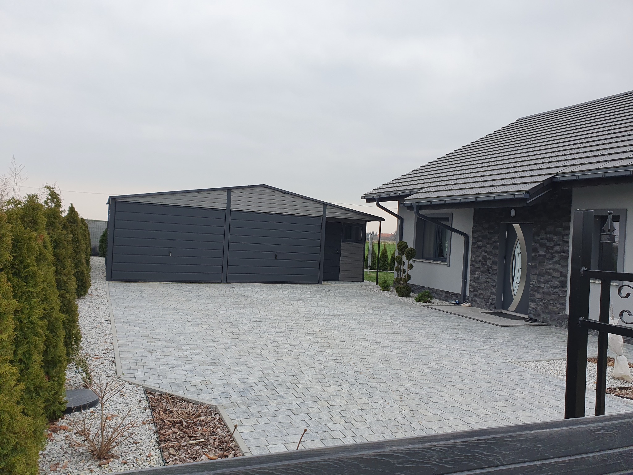 Nowoczesny, dwustanowiskowy garaż z blachy w odcieniach szarości, z bramą segmentową, na posesji z kostką brukową i tujami, widok zza ogrodzenia.