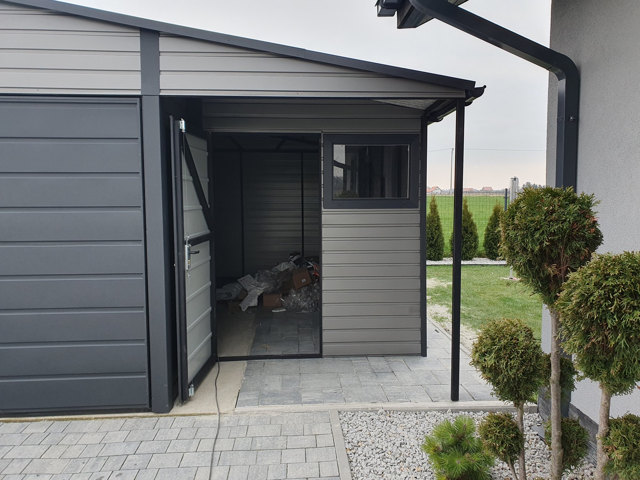 Otwarty, szary garaż blaszany z oknem, widok z zewnątrz na częściowo zagracone wnętrze, kostka brukowa i ozdobne tuje przed garażem.