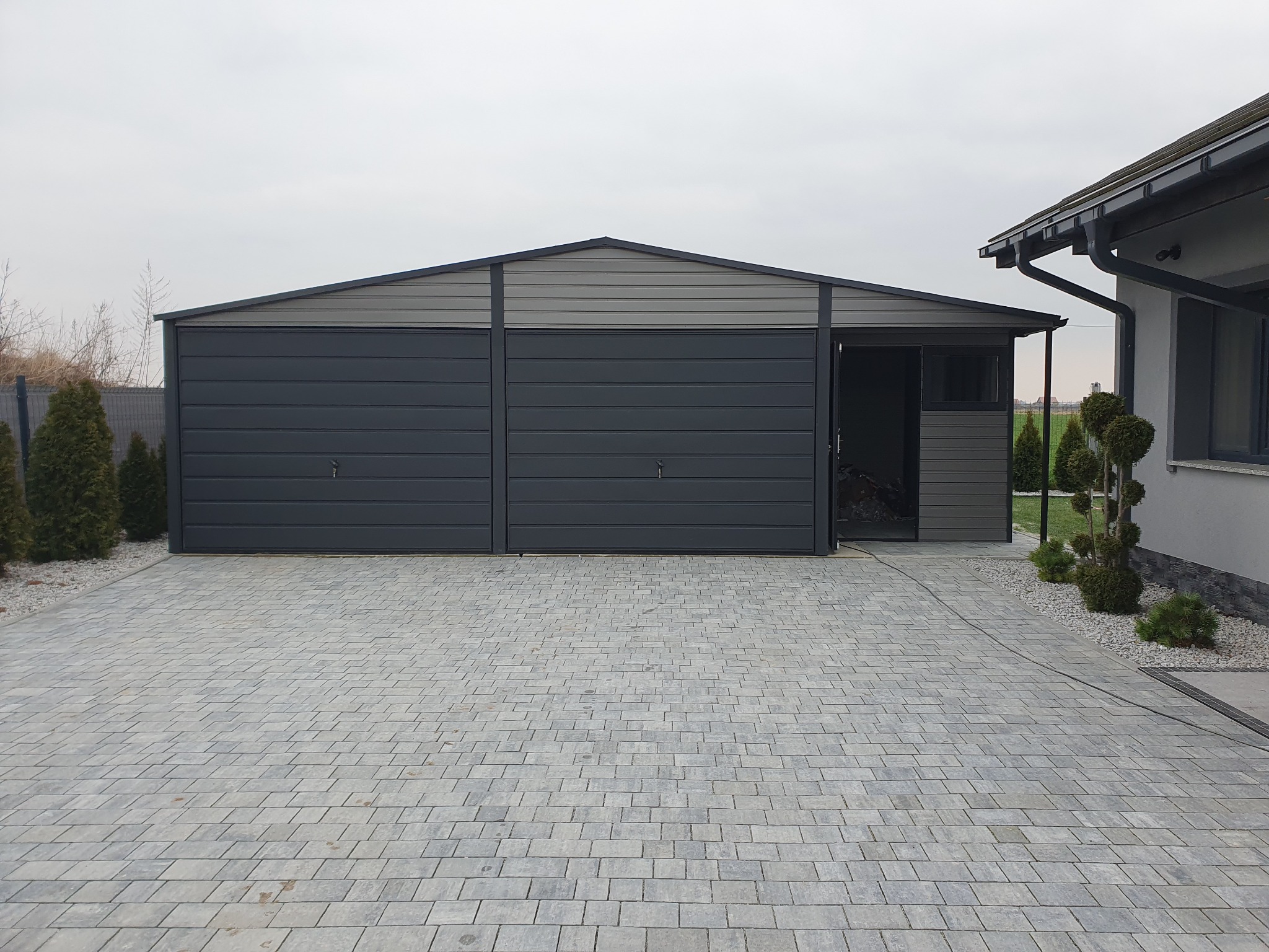 Dwustanowiskowy garaż blaszany w kolorze grafitowym, z bramami segmentowymi i otwartym wejściem bocznym, na posesji z szarą kostką brukową.