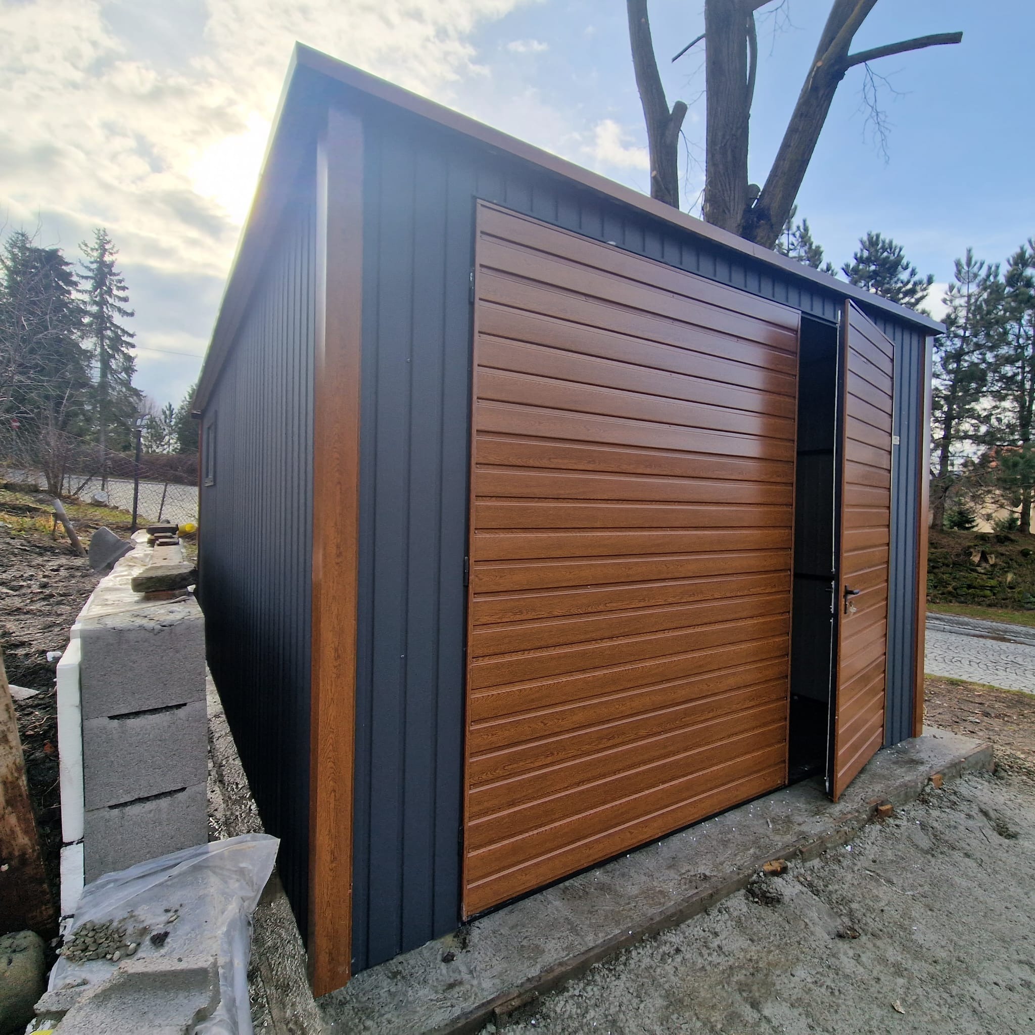 Nowoczesny garaż blaszany z brązową bramą segmentową i szarymi ścianami, częściowo otwarty, widok z boku na tle drzew i nieba.