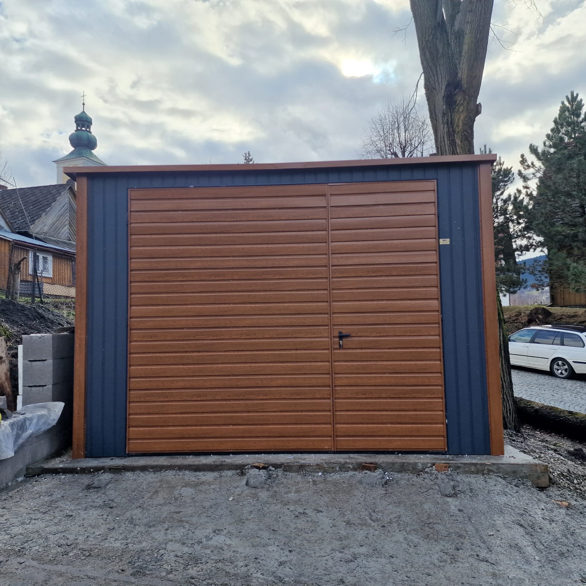 Nowy garaż blaszany z brązową bramą segmentową i grafitowymi ścianami, na betonowej wylewce, w tle wiejski krajobraz z kościołem.