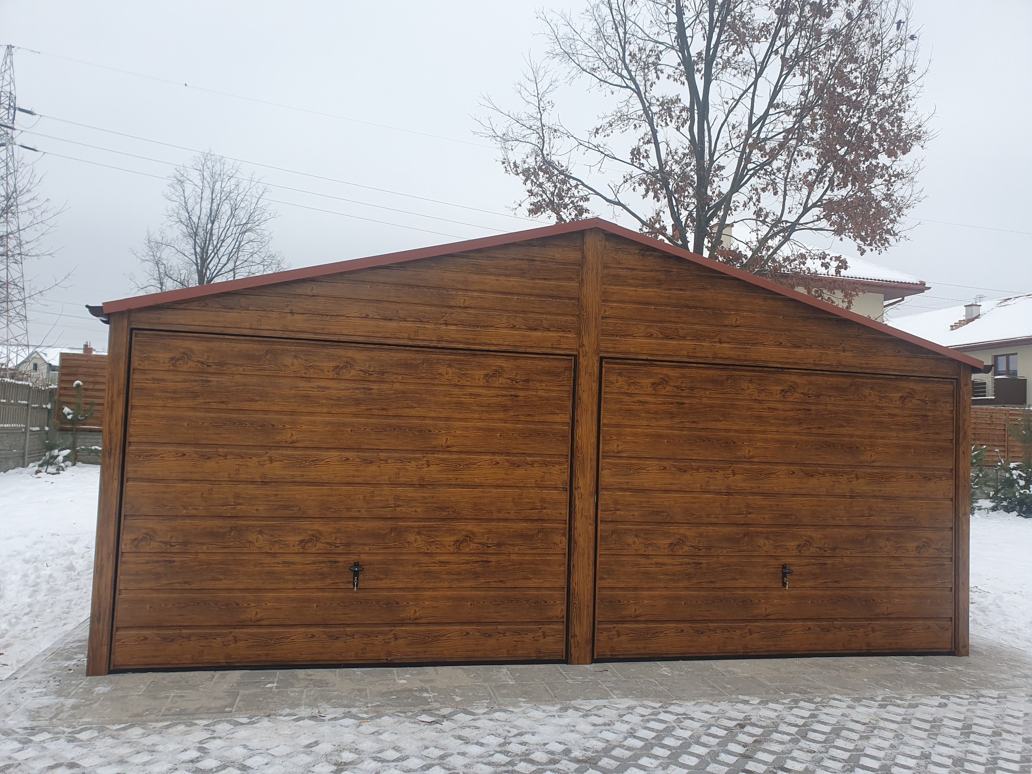 Dwustanowiskowy, drewnopodobny garaż blaszany z dwuspadowym dachem w zimowej scenerii, widoczny na tle zaśnieżonego ogrodu i pochmurnego nieba.