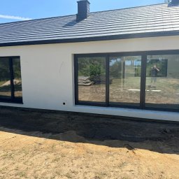 USŁUGI REMONTOWO BUDOWLANE - Nowoczesny dom jednorodzinny z jasną elewacją i dużymi oknami z czarnymi ramami, widoczny fragment dachu z ciemnej dachówki oraz teren wokół budynku w trakcie prac wykończeniowych.