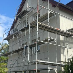USŁUGI REMONTOWO BUDOWLANE - Dom jednorodzinny w trakcie nakładania szarego tynku, widoczne rusztowania oraz fragment dachu z brązową dachówką.